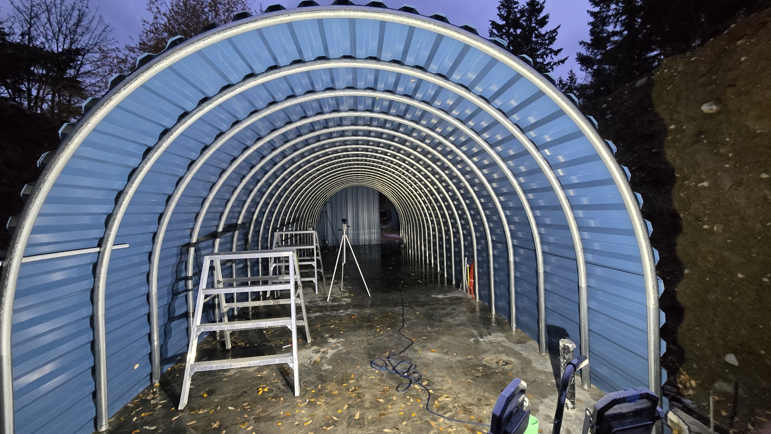 A metal tunnel-like structure with a curved, ribbed roof, illuminated from within. There are ladders and equipment inside, and a tripod with a camera on it. The ground is muddy, and trees are visible around the entrance.