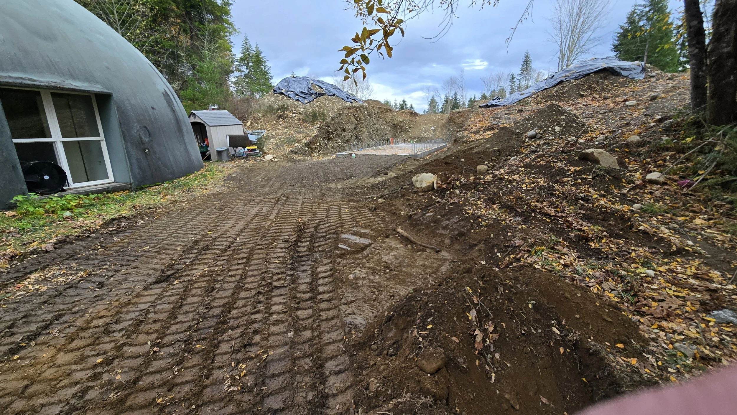 Construction site with disturbed soil, tire tracks on dirt, a rounded building on the left, and trees in the background.