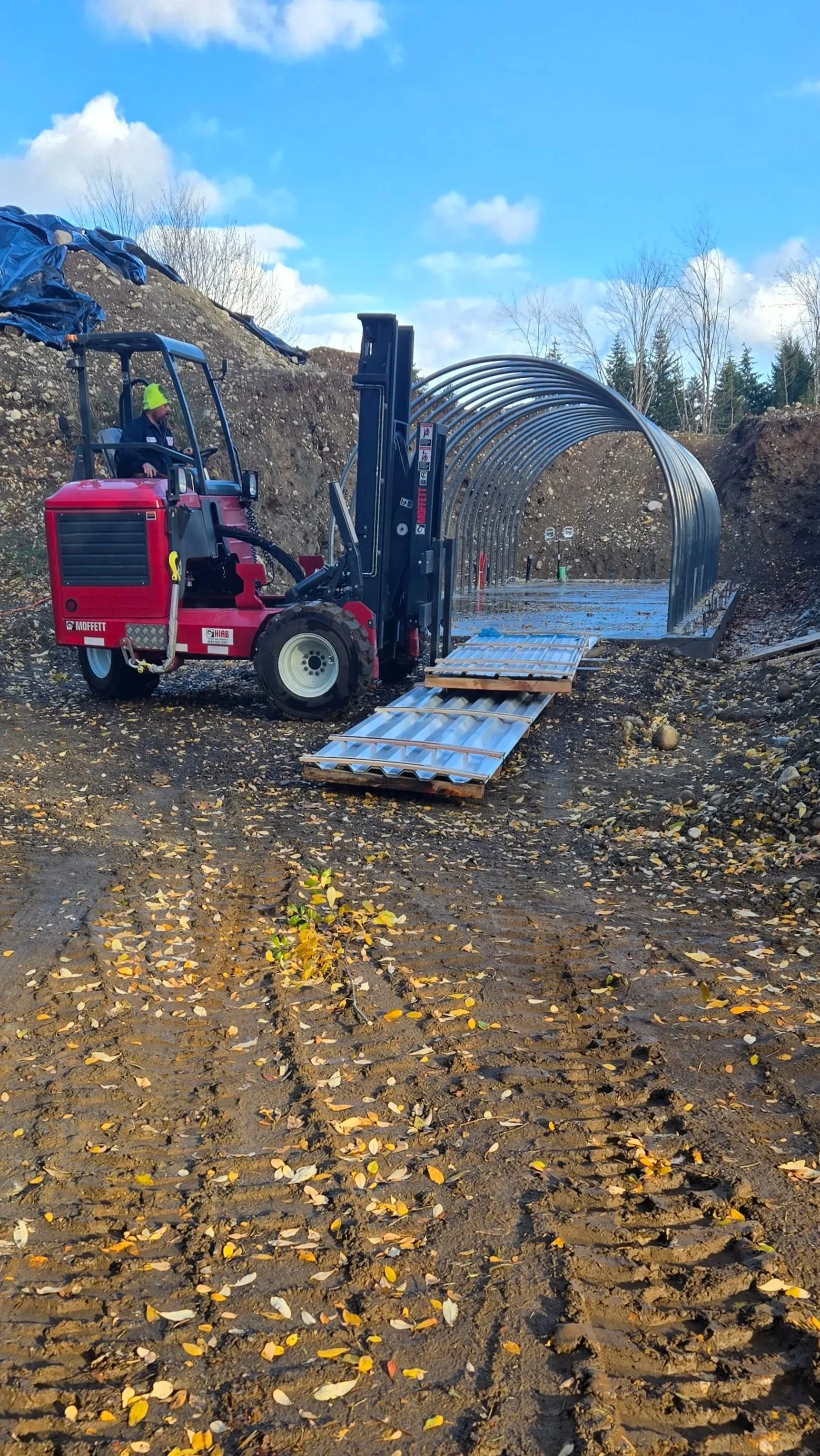 A red forklift moving a metal tunnel structure on a muddy construction site with scattered yellow leaves and a cloudy blue sky.