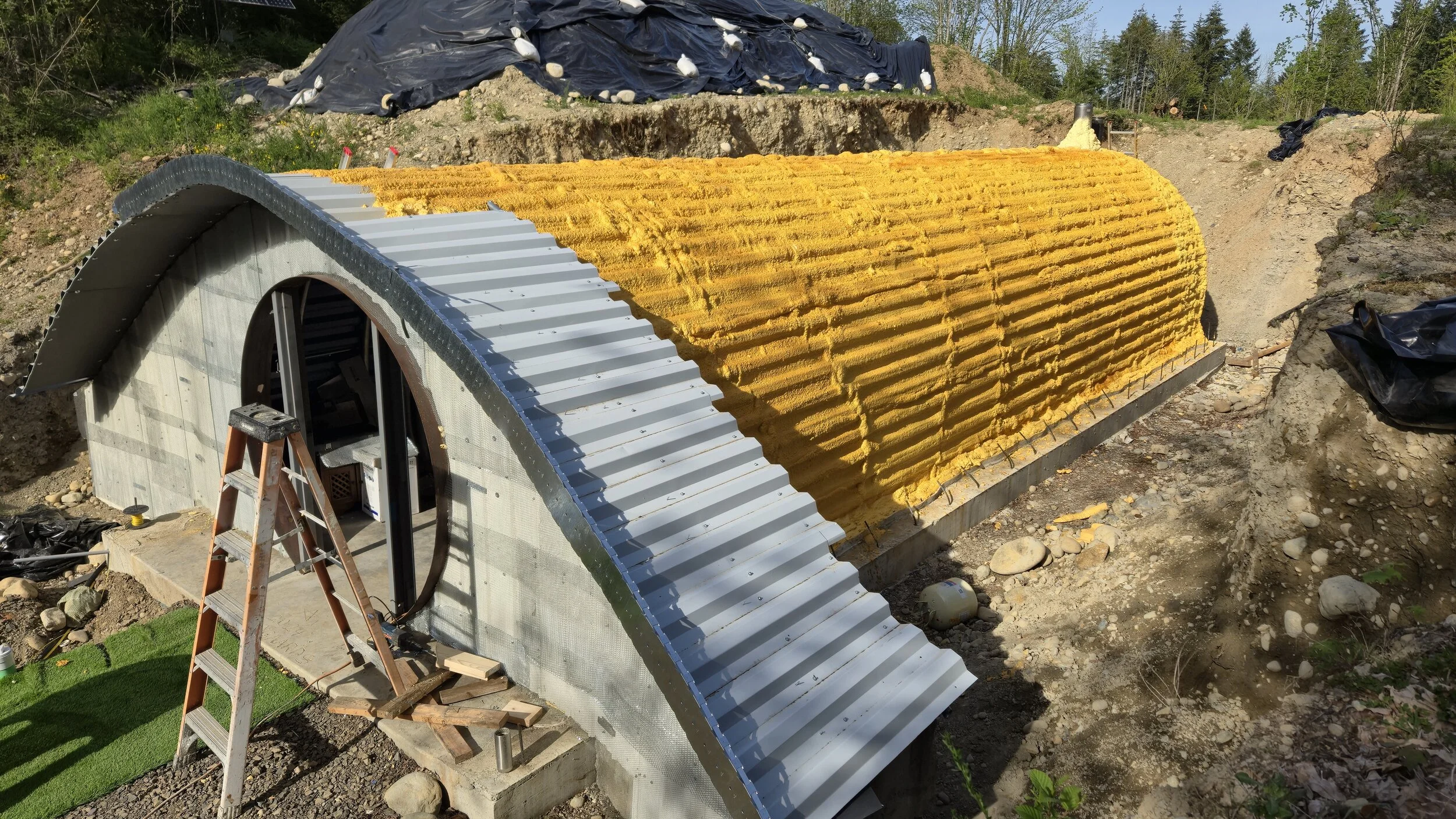 Construction of a small arched underground structure, with concrete and corrugated metal roofing, in a dirt and gravel outdoor setting, with trees in the background.