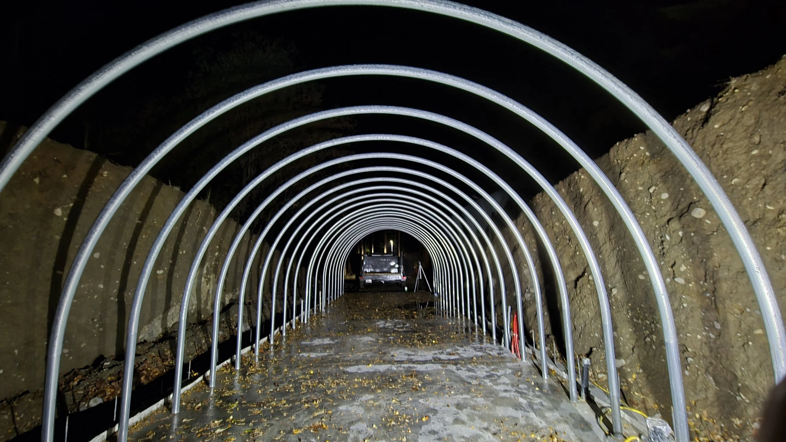 Underground construction site with a metal tunnel frame, dirt walls, a vehicle parked in the distance, and construction materials on the ground at night.