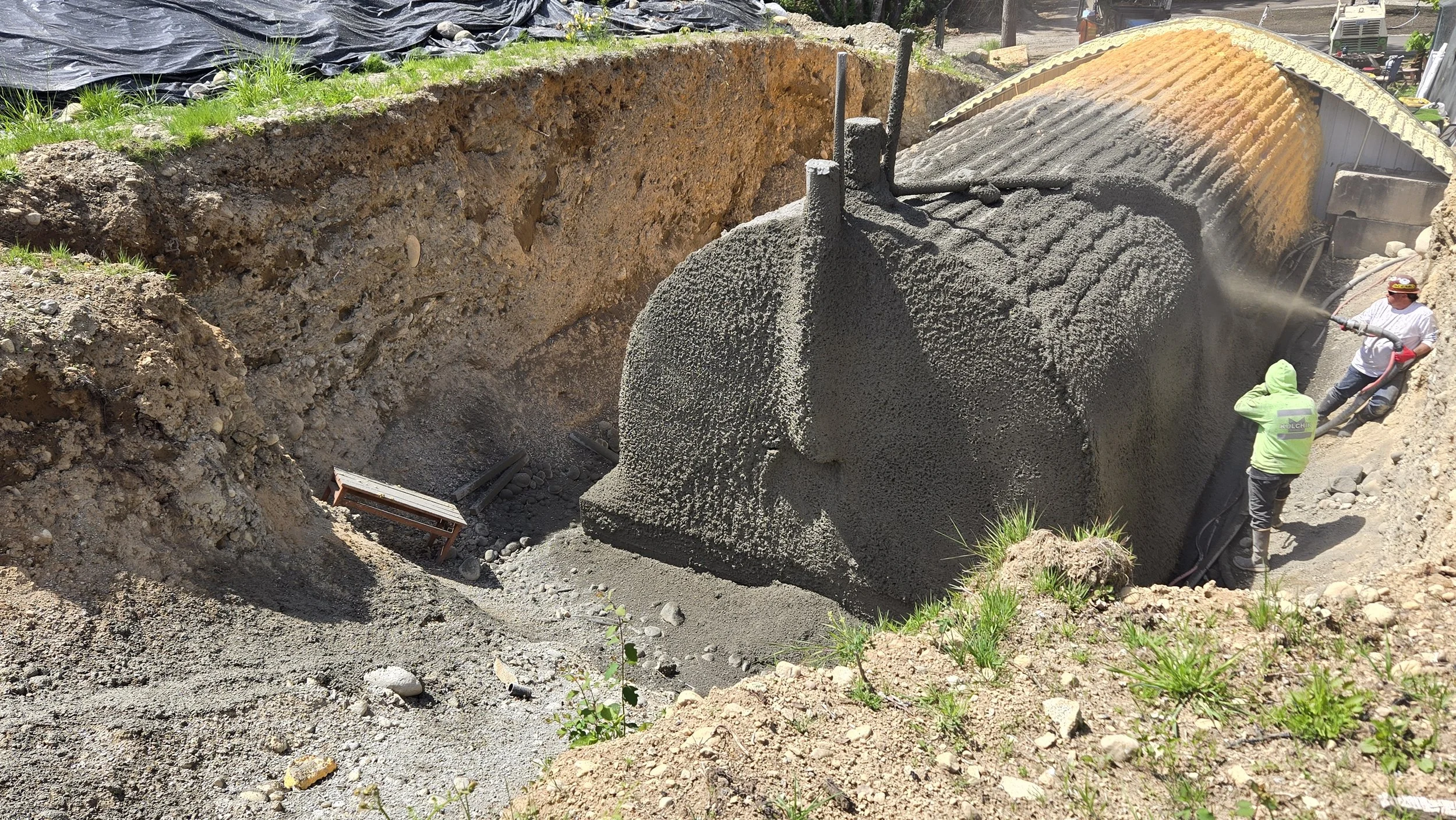Construction workers pouring concrete into a large excavation site with a mix of dirt and gravel, using a truck to deliver the concrete.
