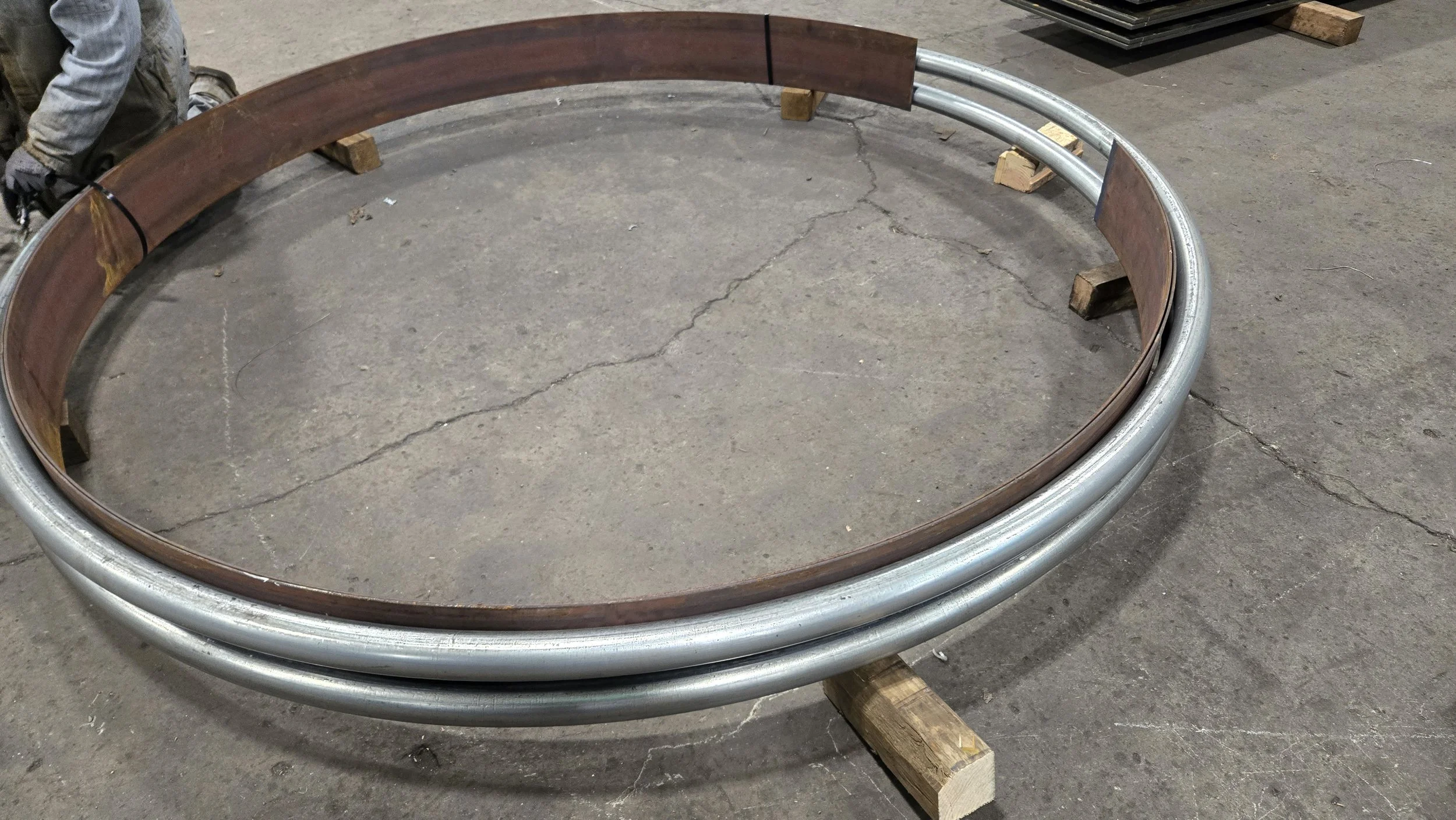 A large circular metal frame with a wooden segment attached, resting on small wooden blocks in a workshop or warehouse setting.