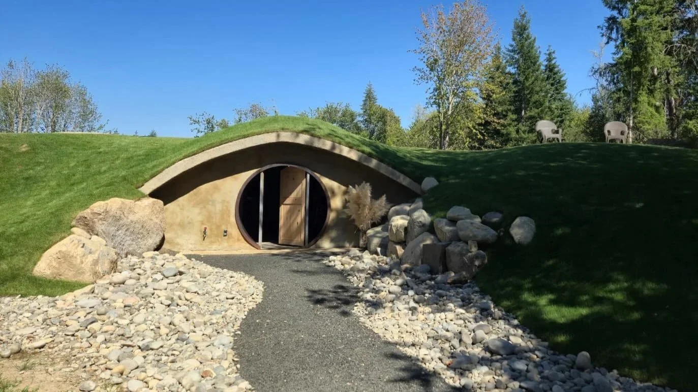 A hobbit-style house with a round door built into a grassy hillside, surrounded by rocks and trees.
