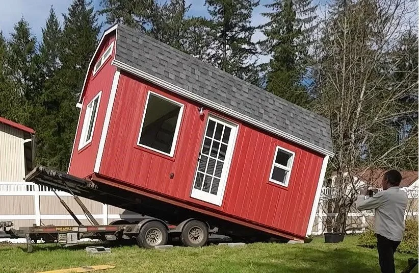 A tiny red house tilted on a trailer during transportation with a man taking a photo nearby.