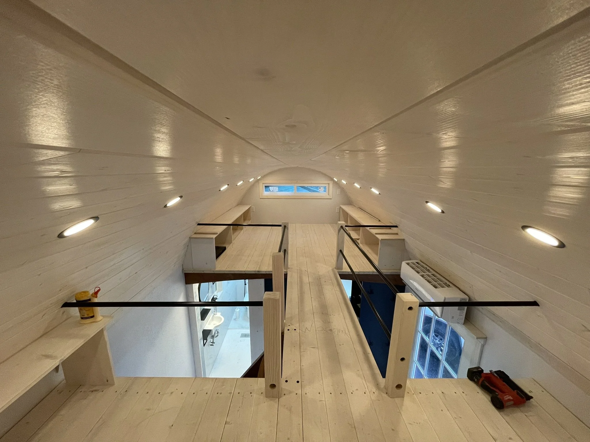 Interior of a house under construction, showing a wooden mezzanine with a safety railing, built-in shelves, and a ceiling with recessed lights. There are windows at the end of the room and construction tools and supplies on the floors.