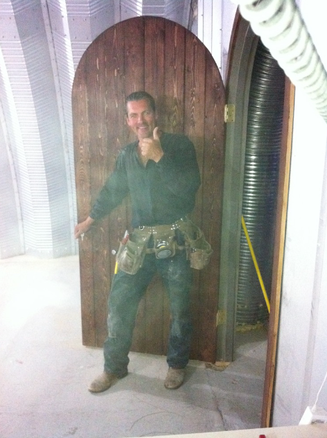 A man in a black shirt and jeans with a tool belt is standing in front of a wooden door, construction or renovation site, with exposed wiring and siding.