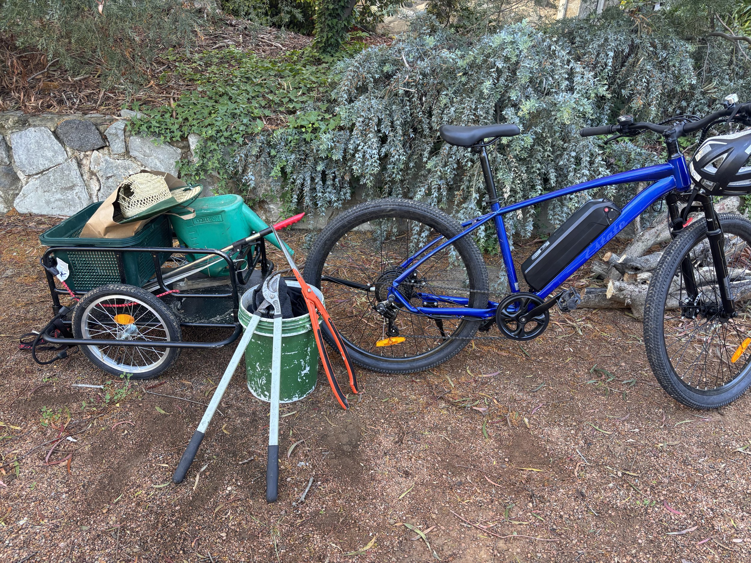 A blue electric mountain bike attached to a black cargo trailer filled with gardening supplies, including a hat, a green watering can, and a garden extra few tools, parked on dirt ground with greenery and rocks in the background.