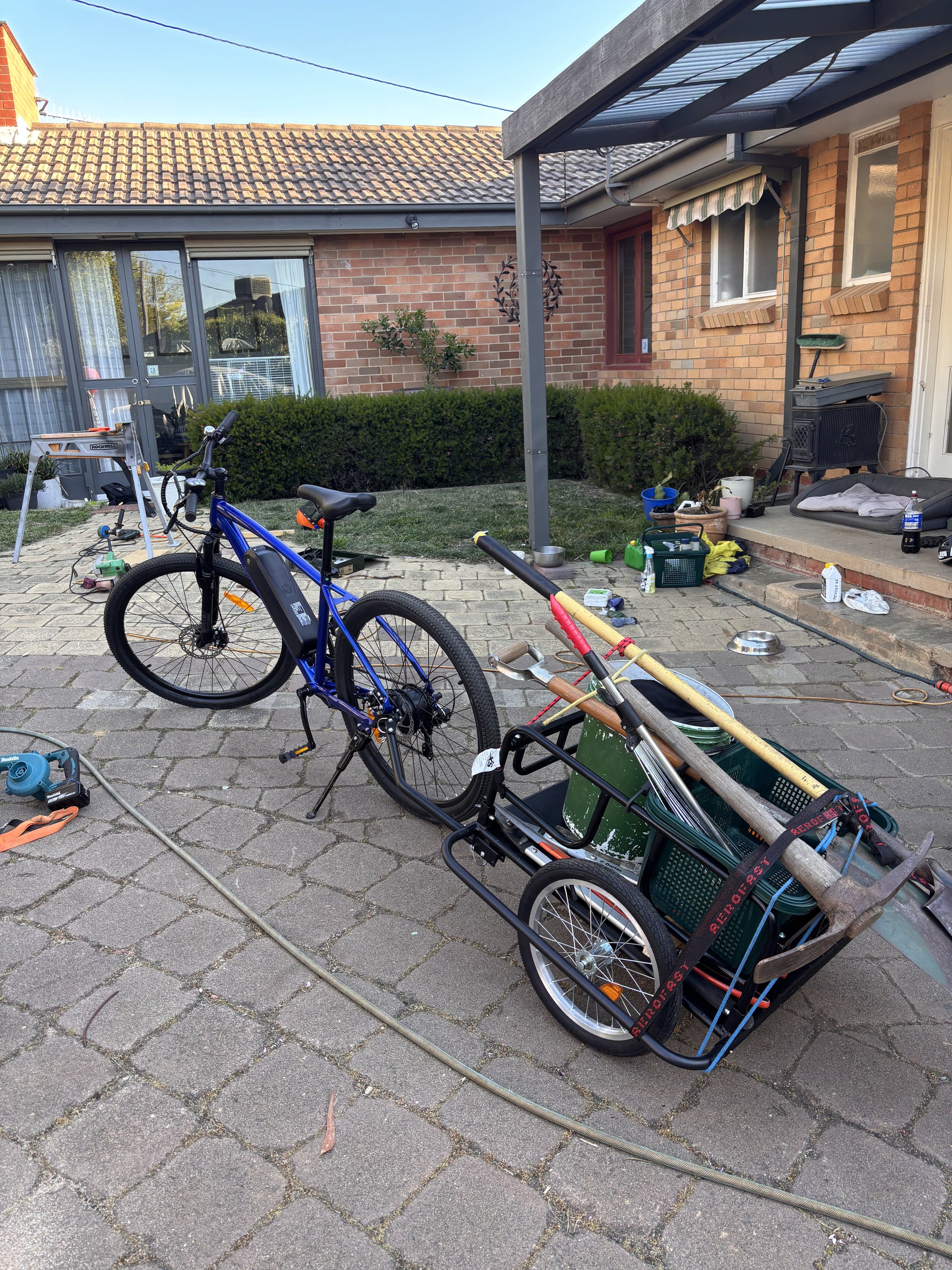 A blue electric bicycle attached to a cargo cart loaded with various tools and equipment, outdoors on a brick patio in front of a house with a brick exterior, small bushes, garden supplies, and outdoor furniture.