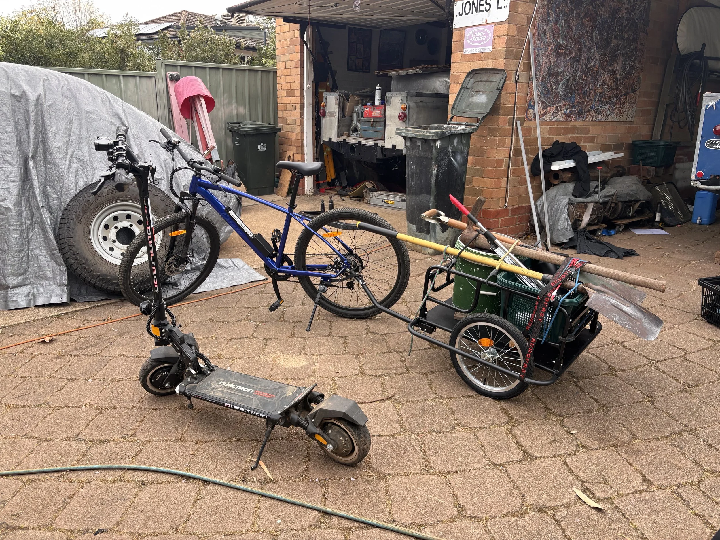A scooter with a flat deck and handlebars, a blue mountain bike, and a cart loaded with gardening tools and supplies are situated on a brick driveway in front of a garage. The garage door is open, revealing tools, parts, and a workbench inside. There