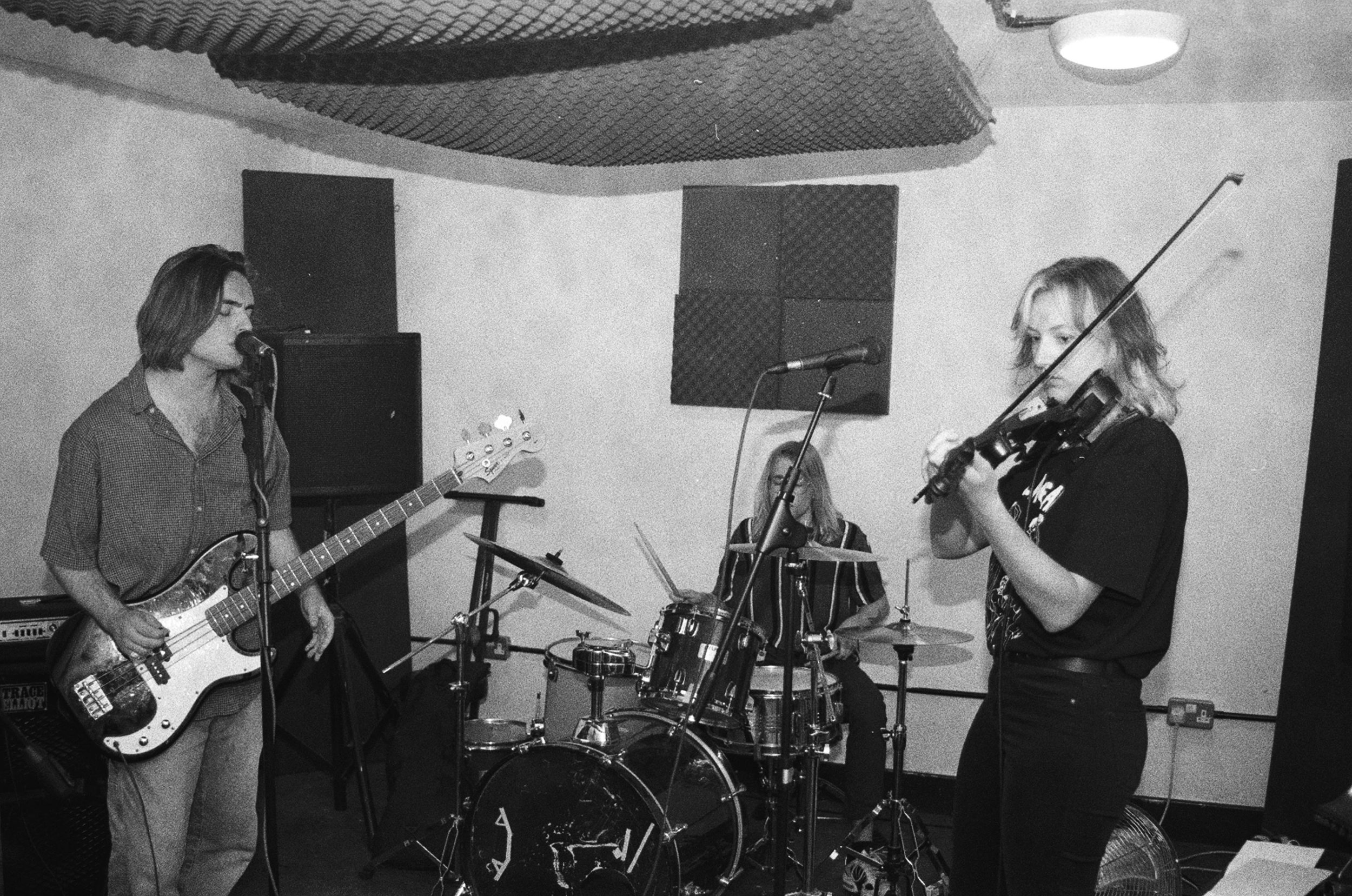 Two men and a woman practicing music in a band, one playing guitar, one on drums, and one playing violin in a music studio.