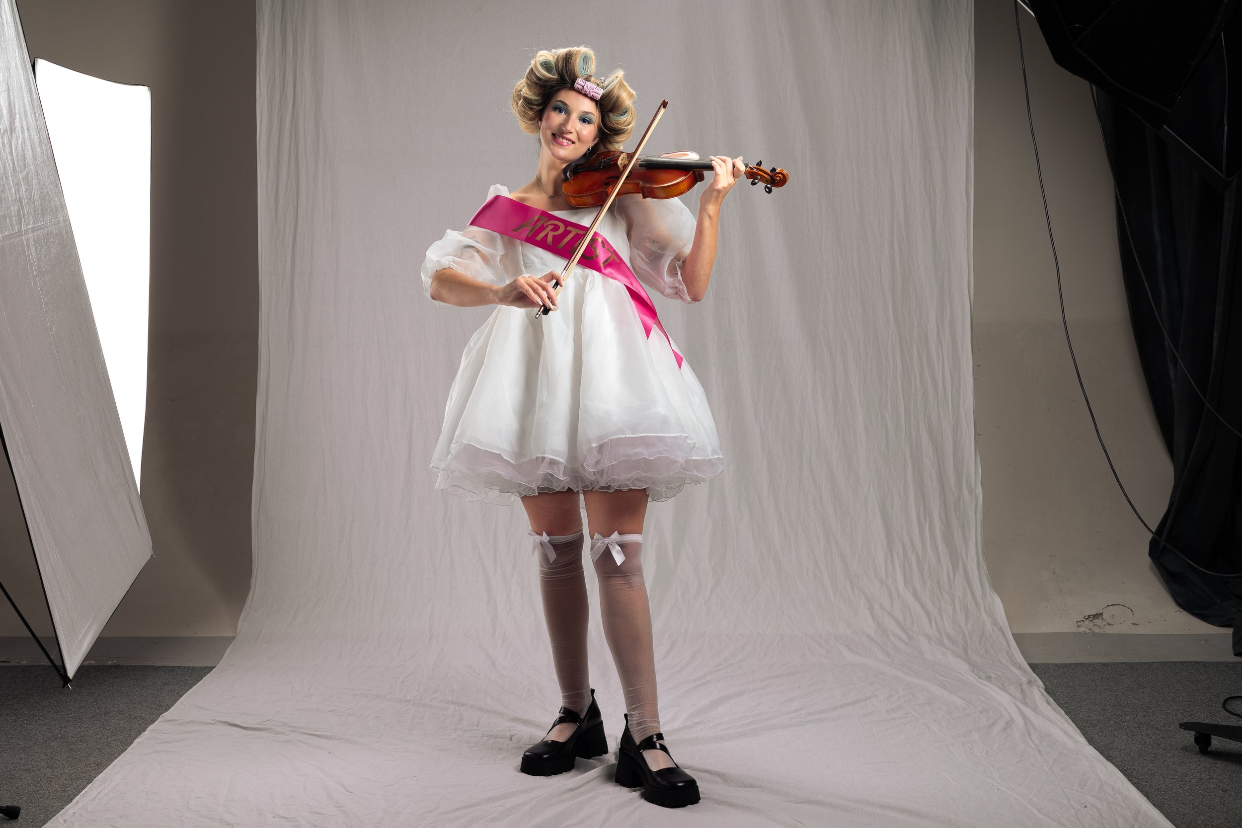 A woman dressed in a white puffy dress with a pink sash that says 'MIRTH', wearing thigh-high stockings with bows, and black platform shoes, is playing a violin in a photography studio with white backdrop and studio lighting.