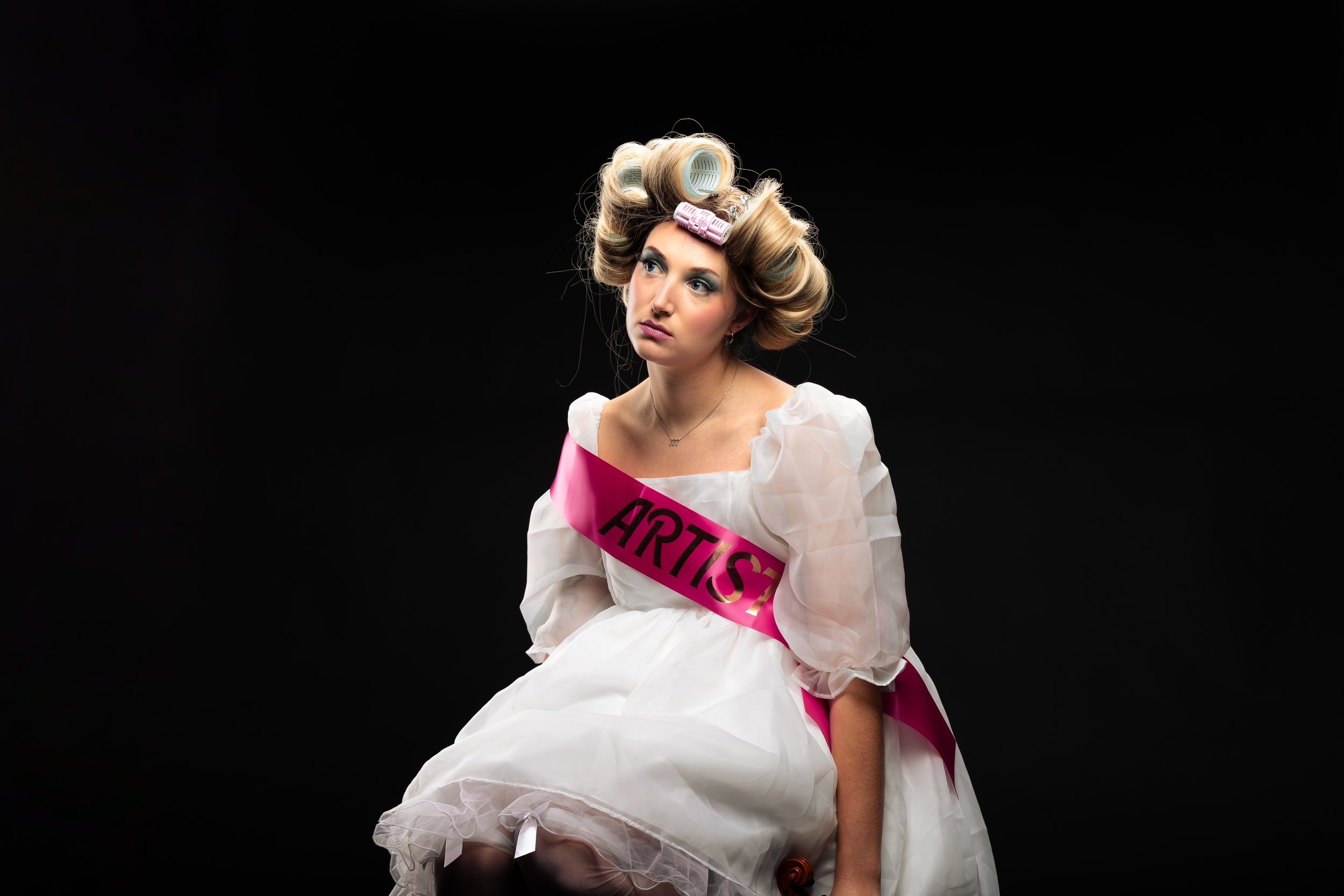 A young woman with blonde hair in curlers and a white dress sits against a black background looking into the distance, wearing a pink sash that reads 'ARTIST'.