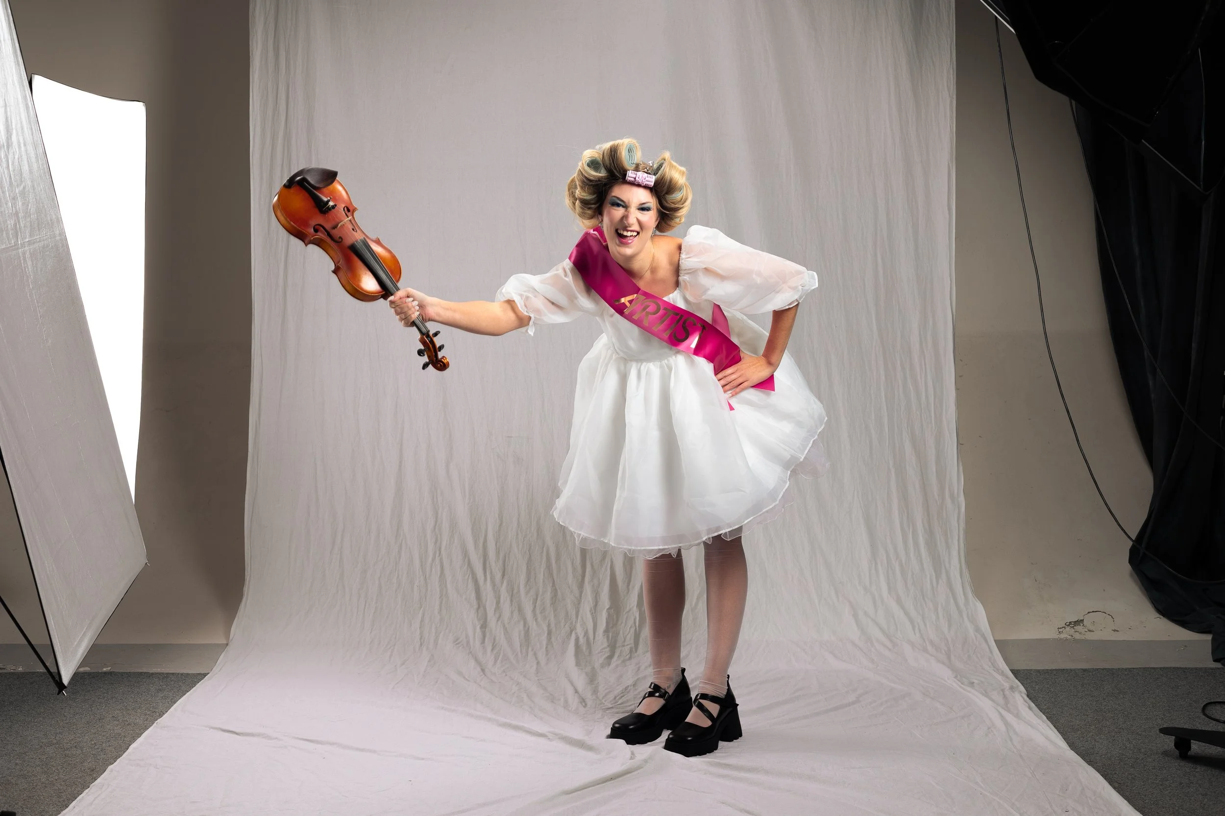 Woman in a white dress and heels about to smash a violin, wearing a pink sash that says "ARTIST," laughing and smiling in a photo studio with white backdrop and lighting equipment.