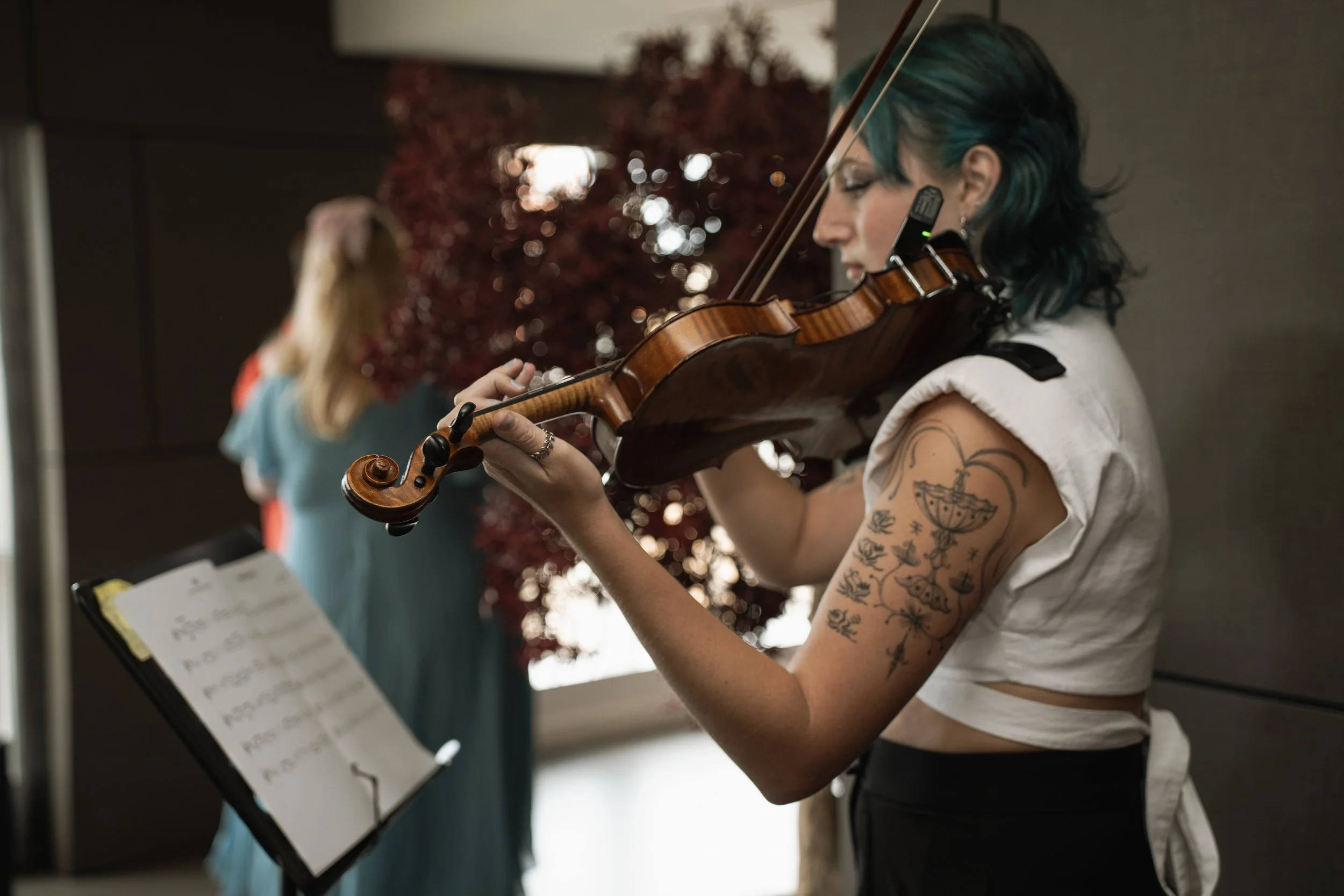 A woman with blue hair and tattoos playing a violin in an indoor setting, with a blurred person in a blue dress in the background.