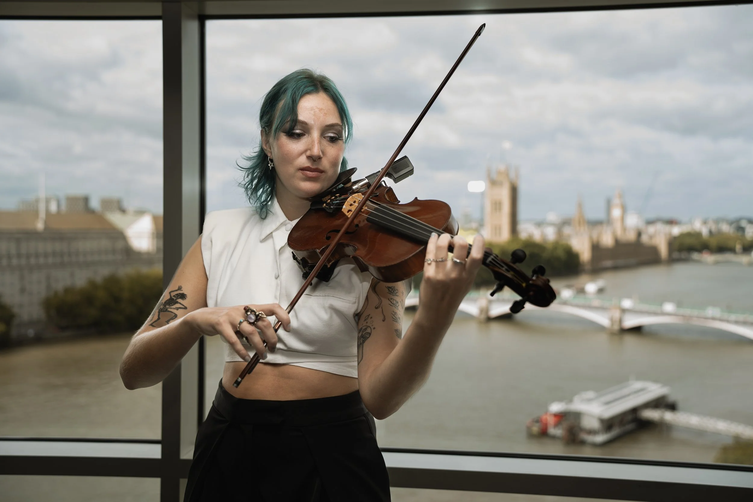 A woman with blue-green hair playing a violin indoors with large windows showing a cityscape of Parliament and the River Thames in the background.