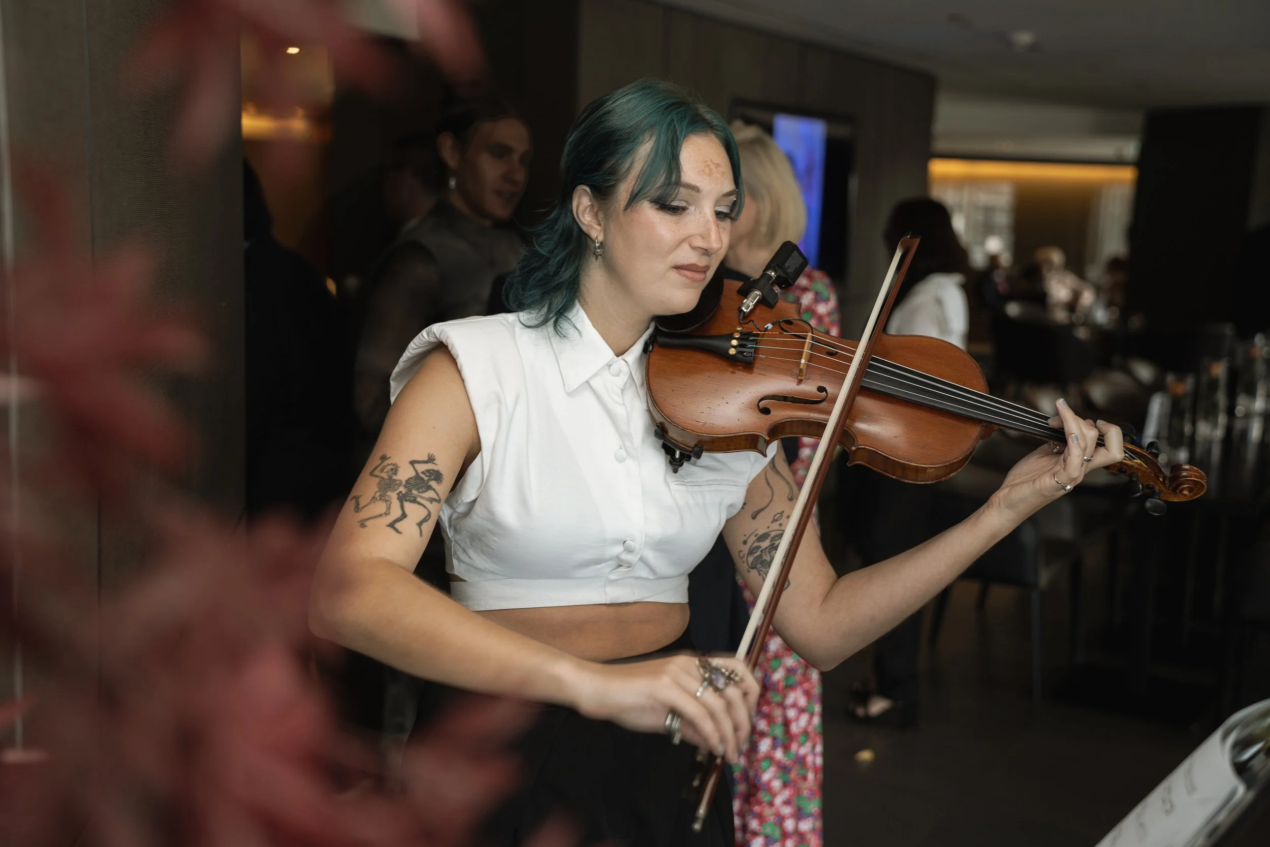 A woman with blue hair and tattoos playing the violin in a lively indoor setting.