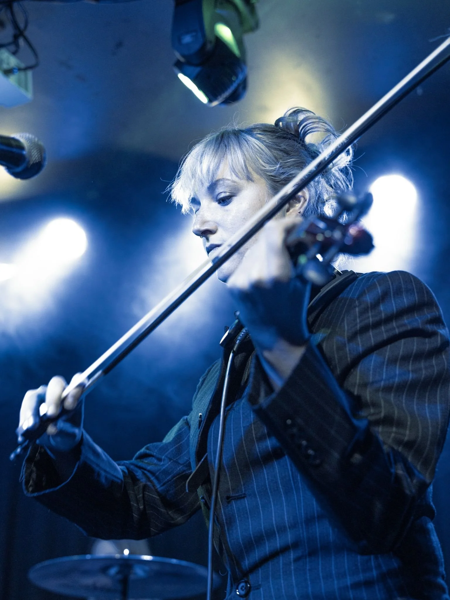 A female musician playing the violin on stage under blue lighting, wearing a pinstripe suit