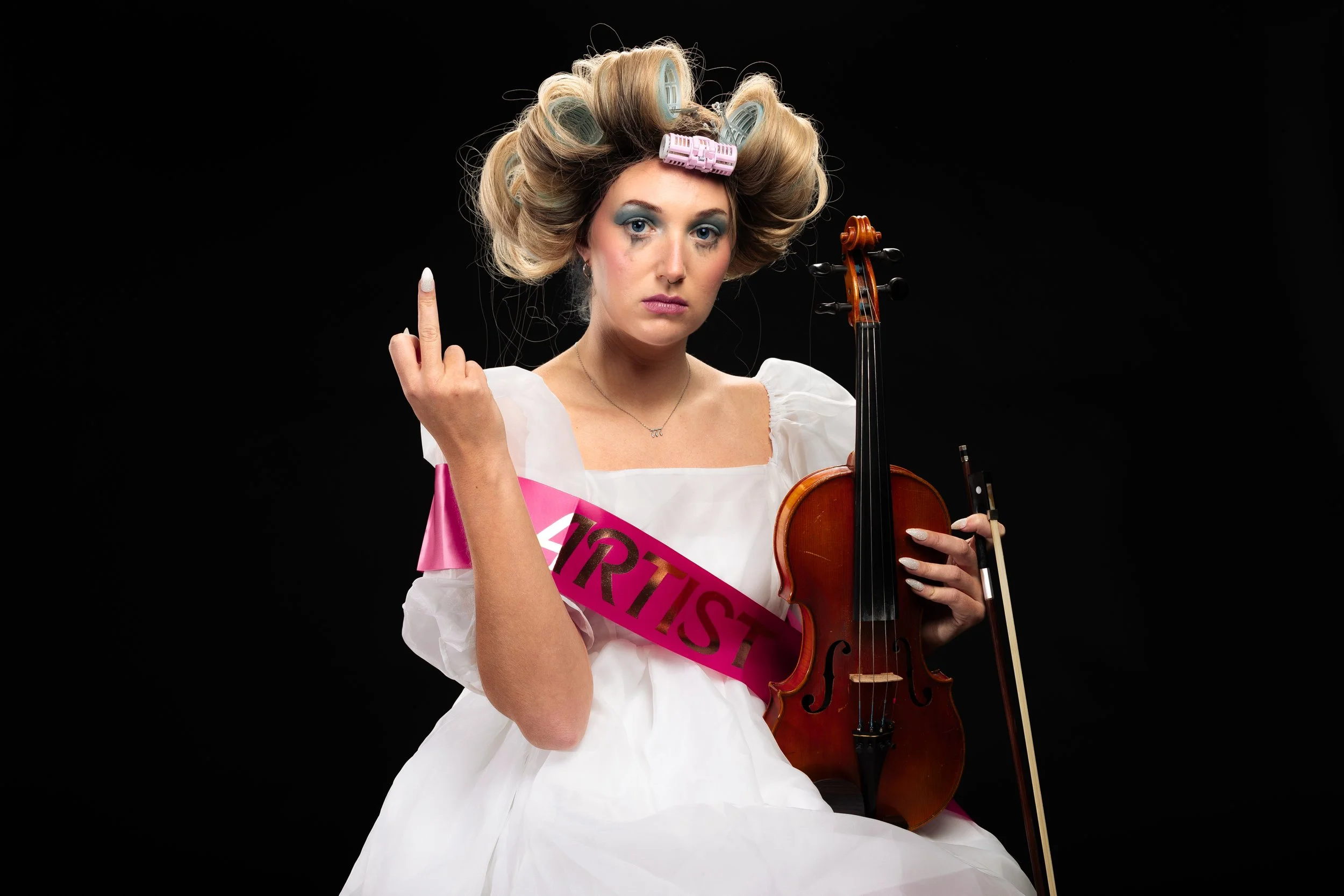 A woman with curlers in her hair, wearing a white dress and a pink sash that says 'ARTIST,' holding a violin and bow, and making an obscene gesture with her middle finger against a black background.