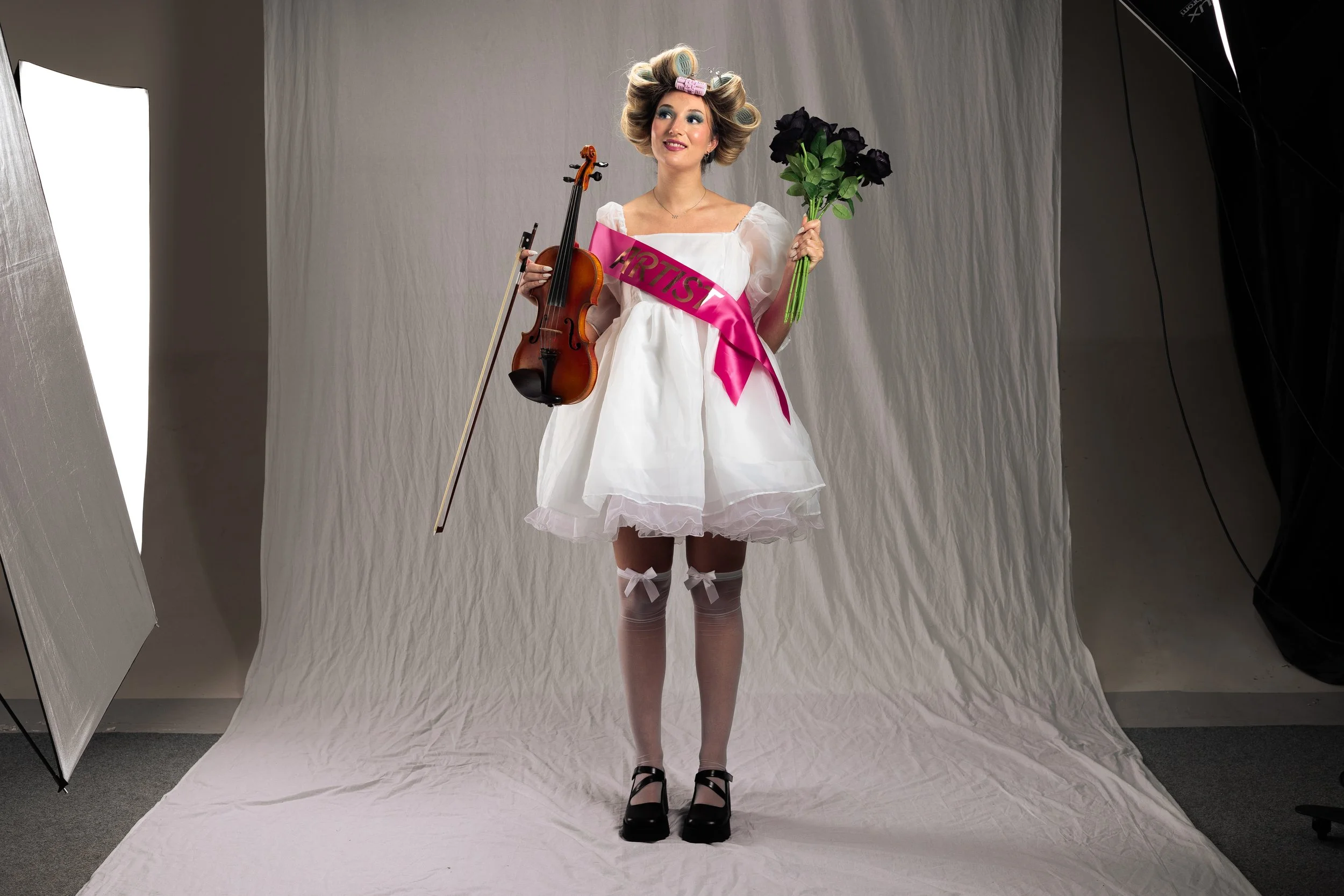 Young woman in a white dress with puffy sleeves, pink sash labeled 'ARTIST,' holding a violin and a bouquet of black roses, standing on a white backdrop in a photo studio.