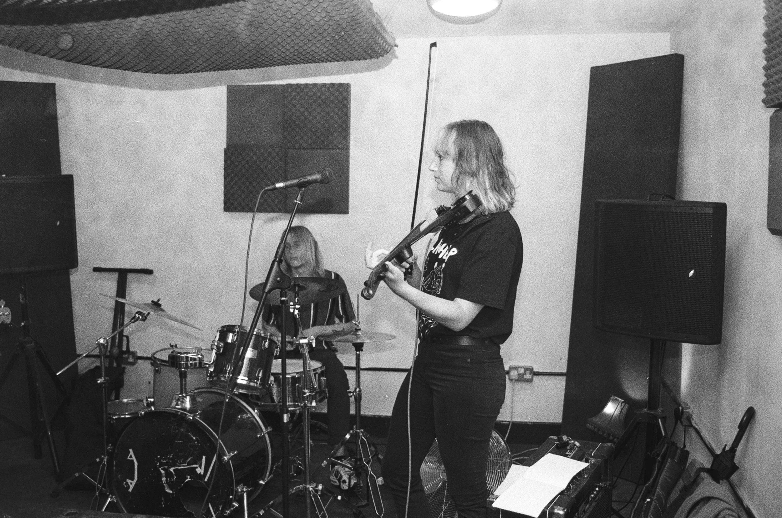 A man and a woman performing music in a recording studio: one playing drums and the other playing a violin.
