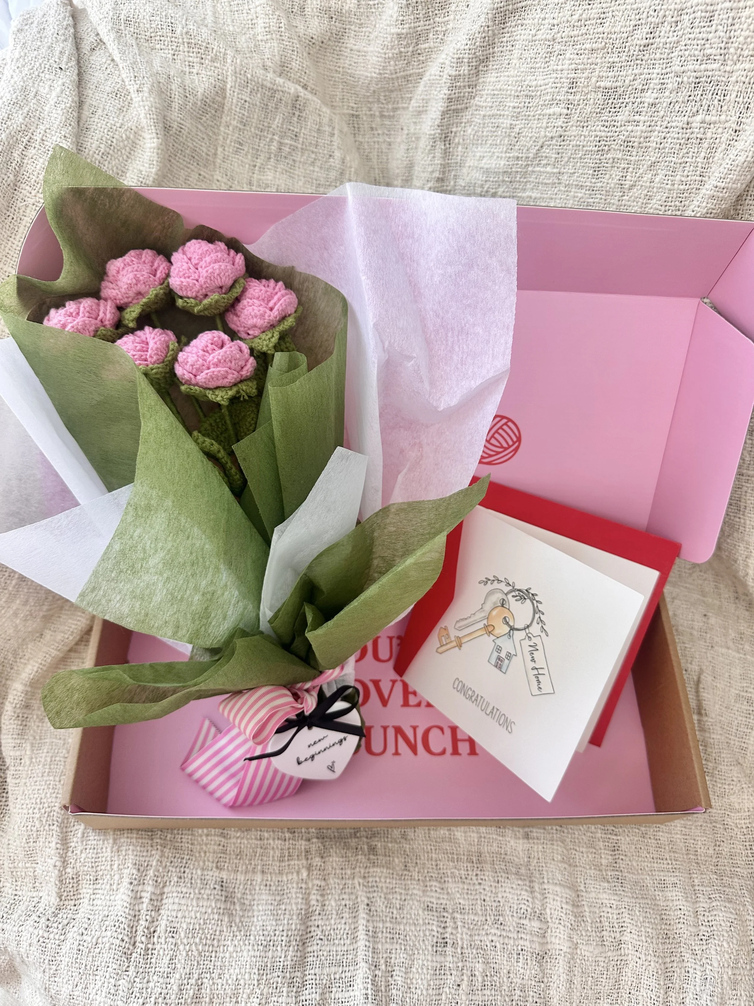 A pink gift box contains a bouquet of pink crochet flowers, a congratulatory card with a key illustration, and a small wrapped item with a pink and white striped ribbon.