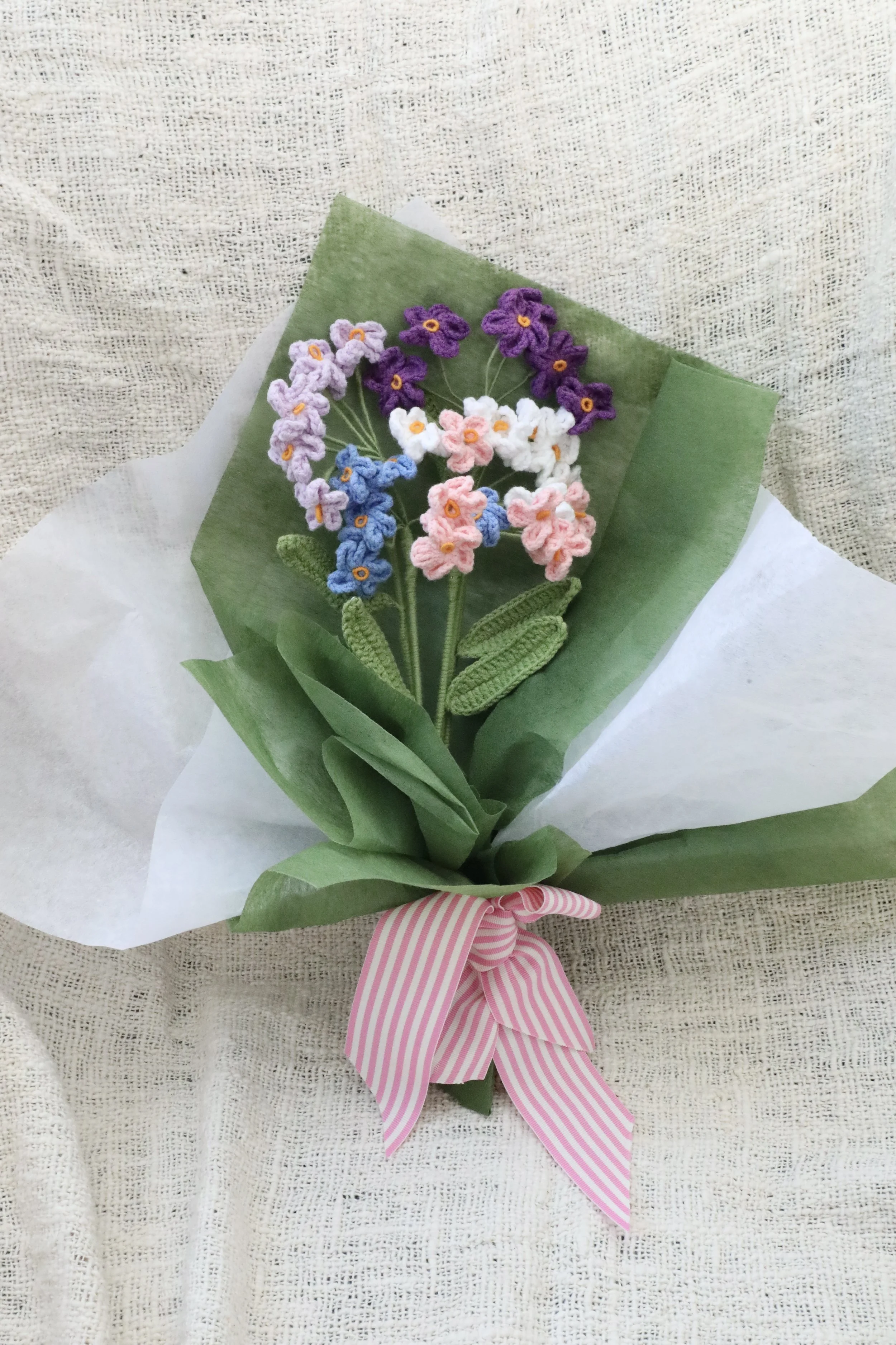 A bouquet of crochet flowers with pink, purple, white, and blue Forget Me Not blooms, wrapped in green and white tissue paper, and tied with a pink and white striped ribbon.