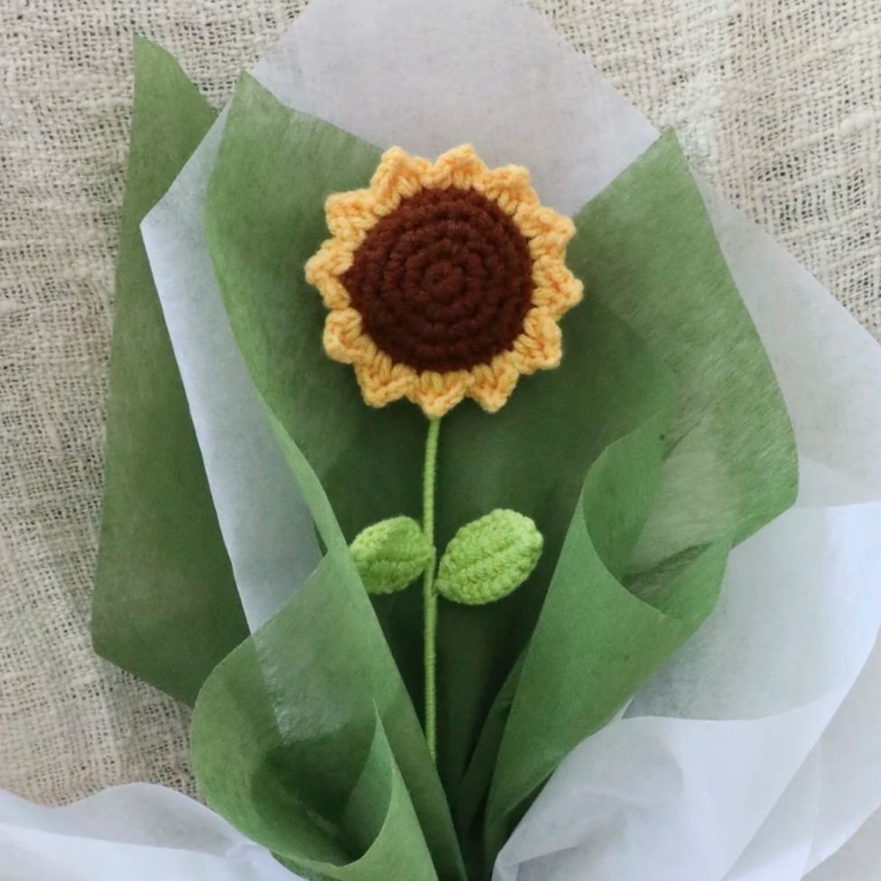 A crocheted sunflower with yellow petals, a brown center, and two green leaves, wrapped in green and white paper.