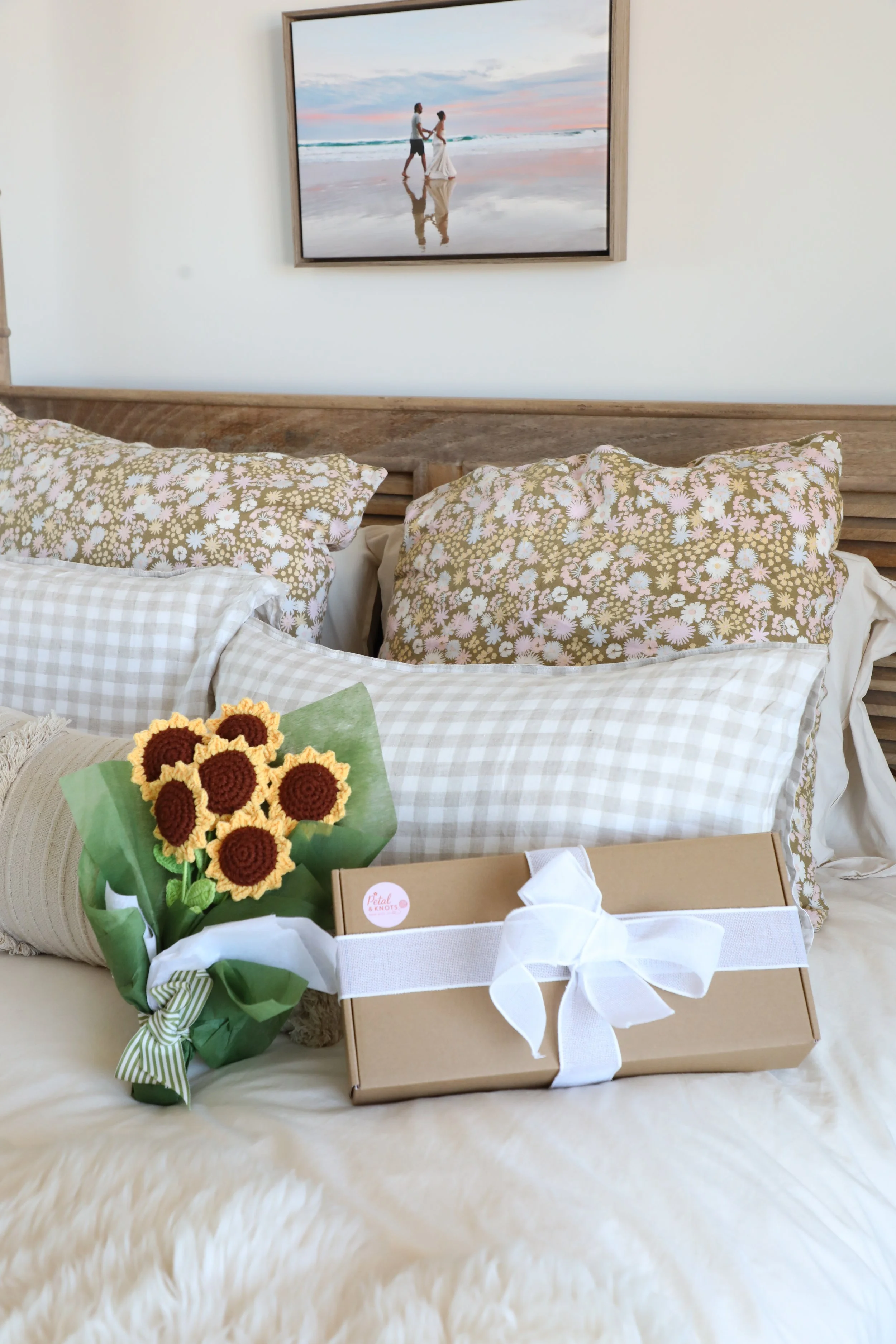 A bed with floral and gingham pillows, a bouquet of crochet sunflowers, and a gift box with a white ribbon in a bedroom, with a framed beach photograph hanging on the wall above.