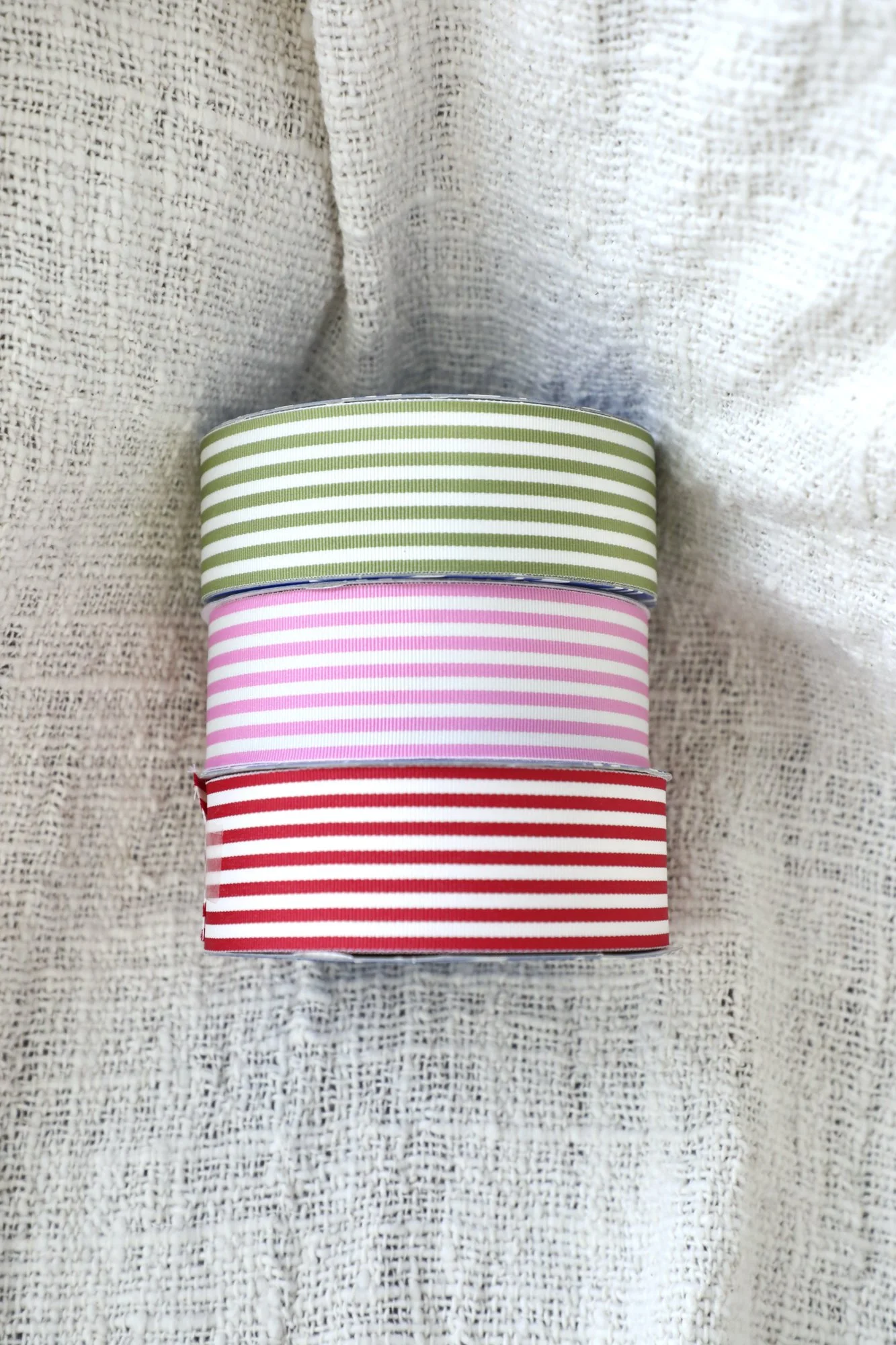 Three stacked spools of ribbon in green, pink, and red with white stripes, placed on a textured beige fabric background.