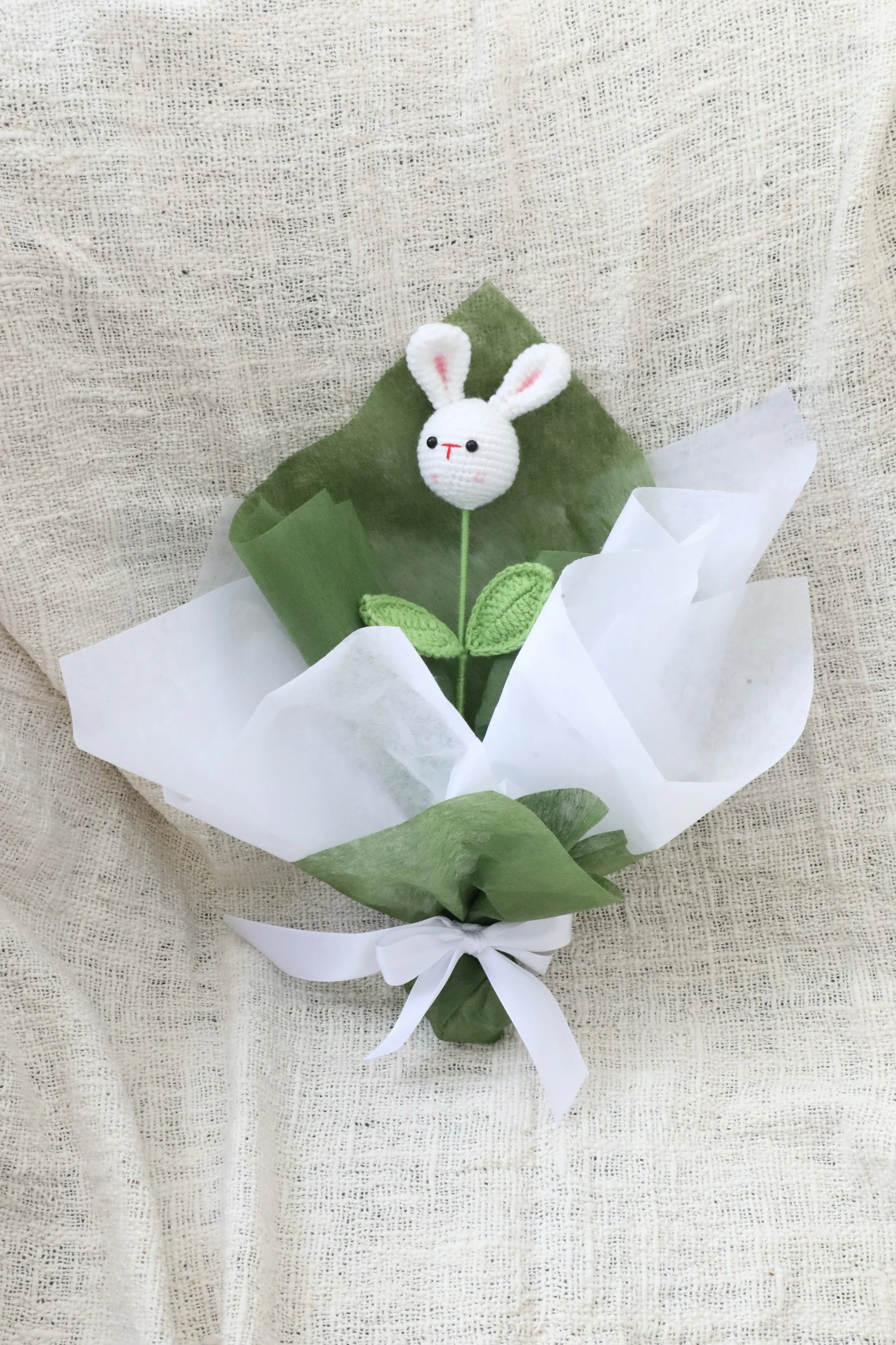 A decorative crochet bunny on a bouquet of green leaves wrapped in white tissue paper and tied with a white ribbon.