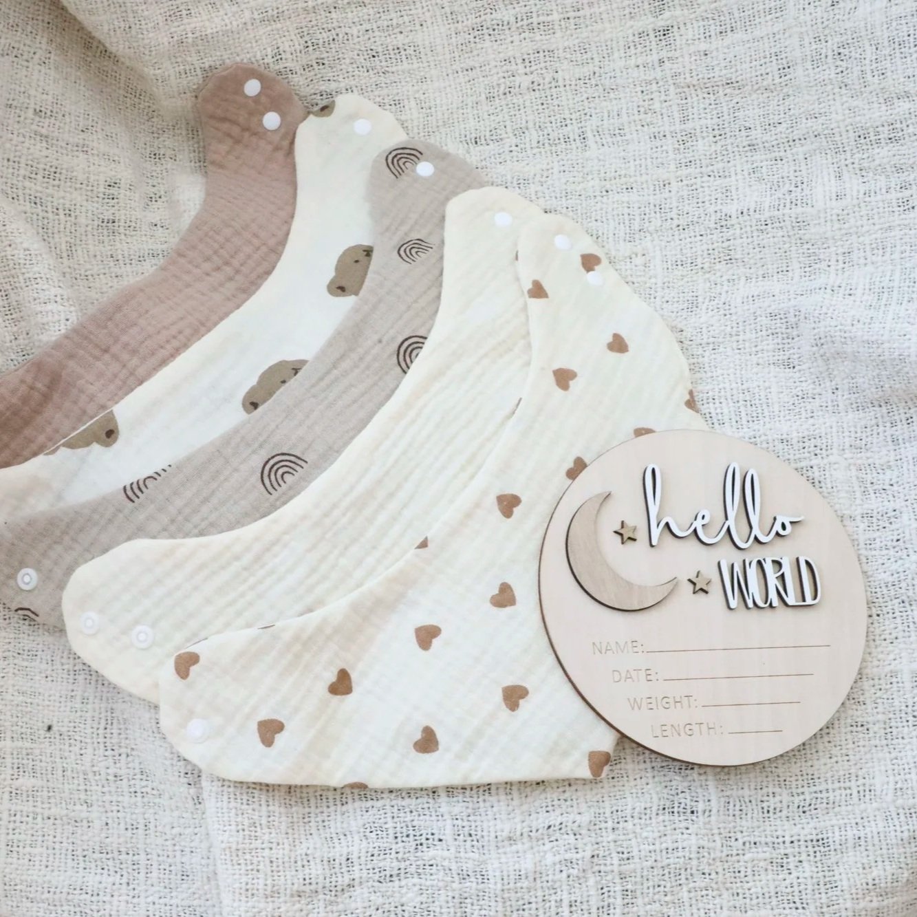 Four patterned baby bibs laid out on a textured fabric surface with a circular wooden plaque that reads 'hello WORLD' and has spaces for baby’s name, date, weight, and length.
