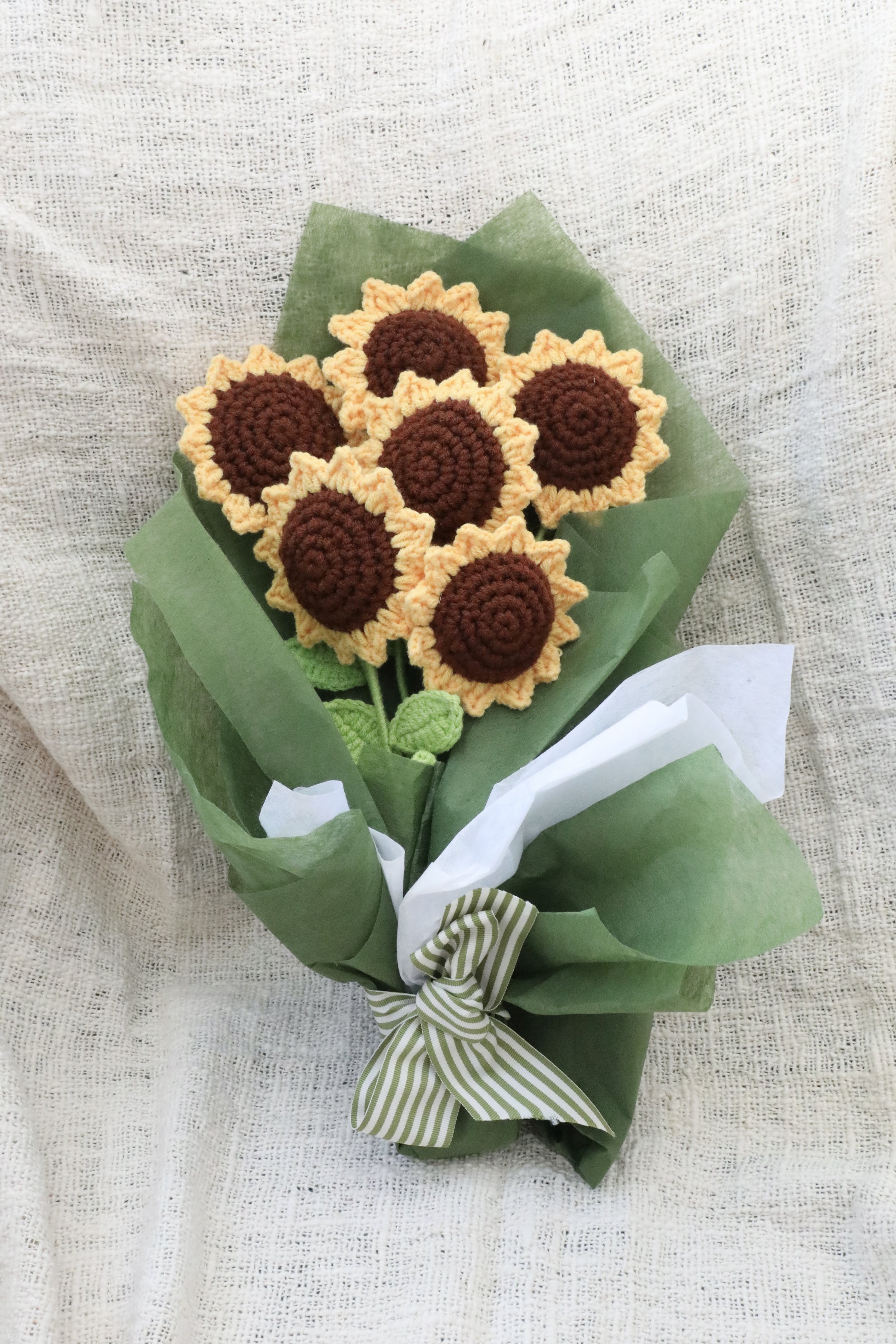 A bouquet of handcrafted sunflower crochet flowers wrapped in green and white paper, tied with a green and white striped ribbon.