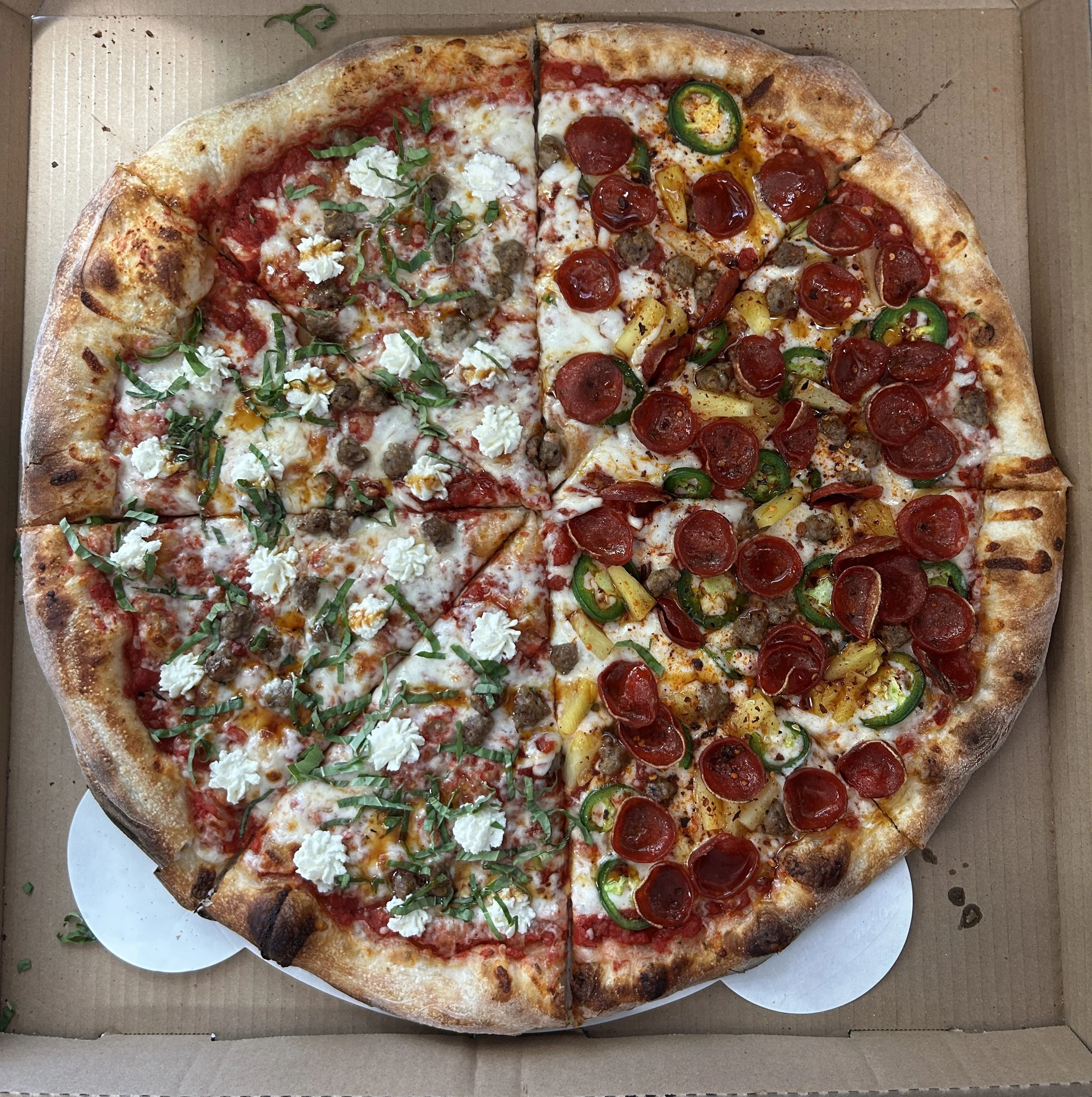 A large pizza with half topped with cheese, sausage, green herbs, and white crumbles, and the other half with pepperoni, green peppers, and yellow peppers, in a pizza box