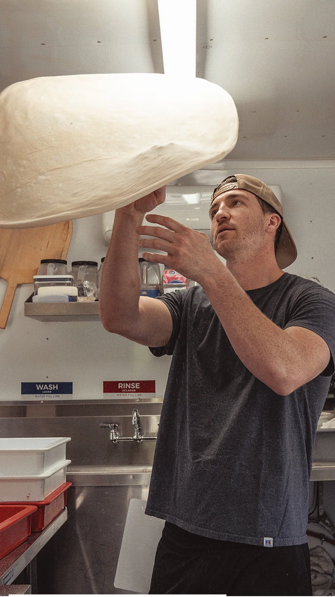 A man in a black t-shirt and a backward baseball cap is tossing pizza dough in the air in a commercial kitchen.