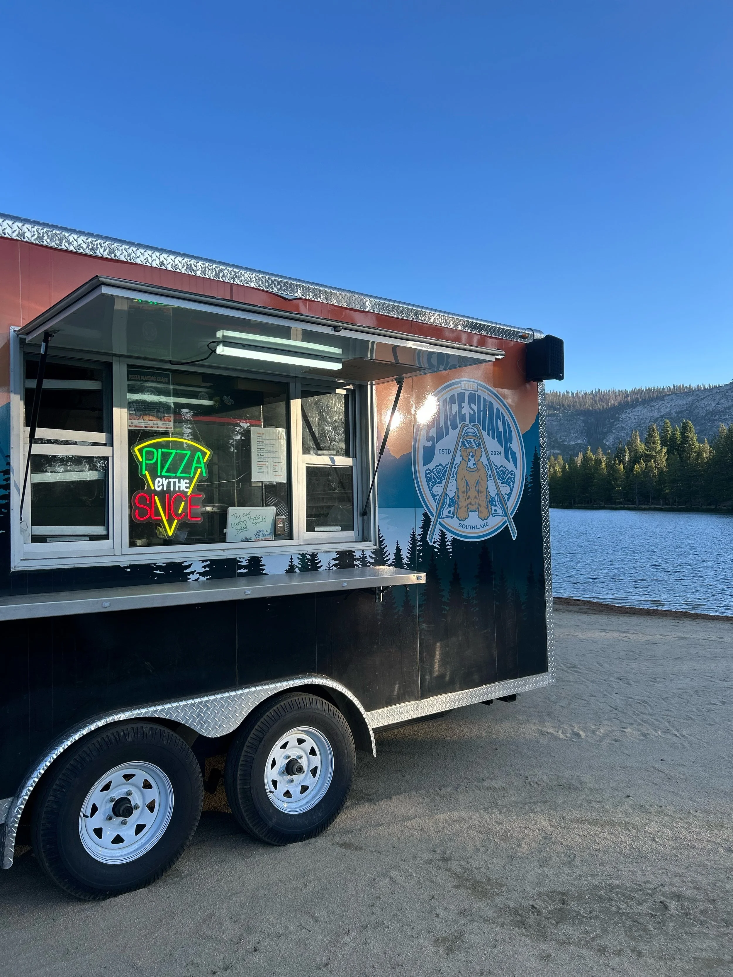 A food truck parked beside a lake with trees and mountains in the background. The truck features a logo with a bear and the words 'The SteSHACK ESTD 2024 South Lake.' The window displays a neon sign that reads 'PIZZA THE' in green, 'SLICE' in red, an