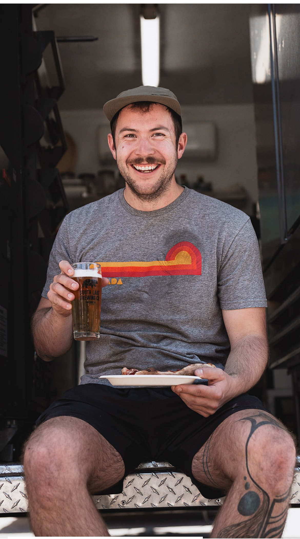 A smiling man sitting inside a food truck, holding a pint of beer in one hand and a plate with food in the other, wearing a gray T-shirt, black shorts, and a beige cap.