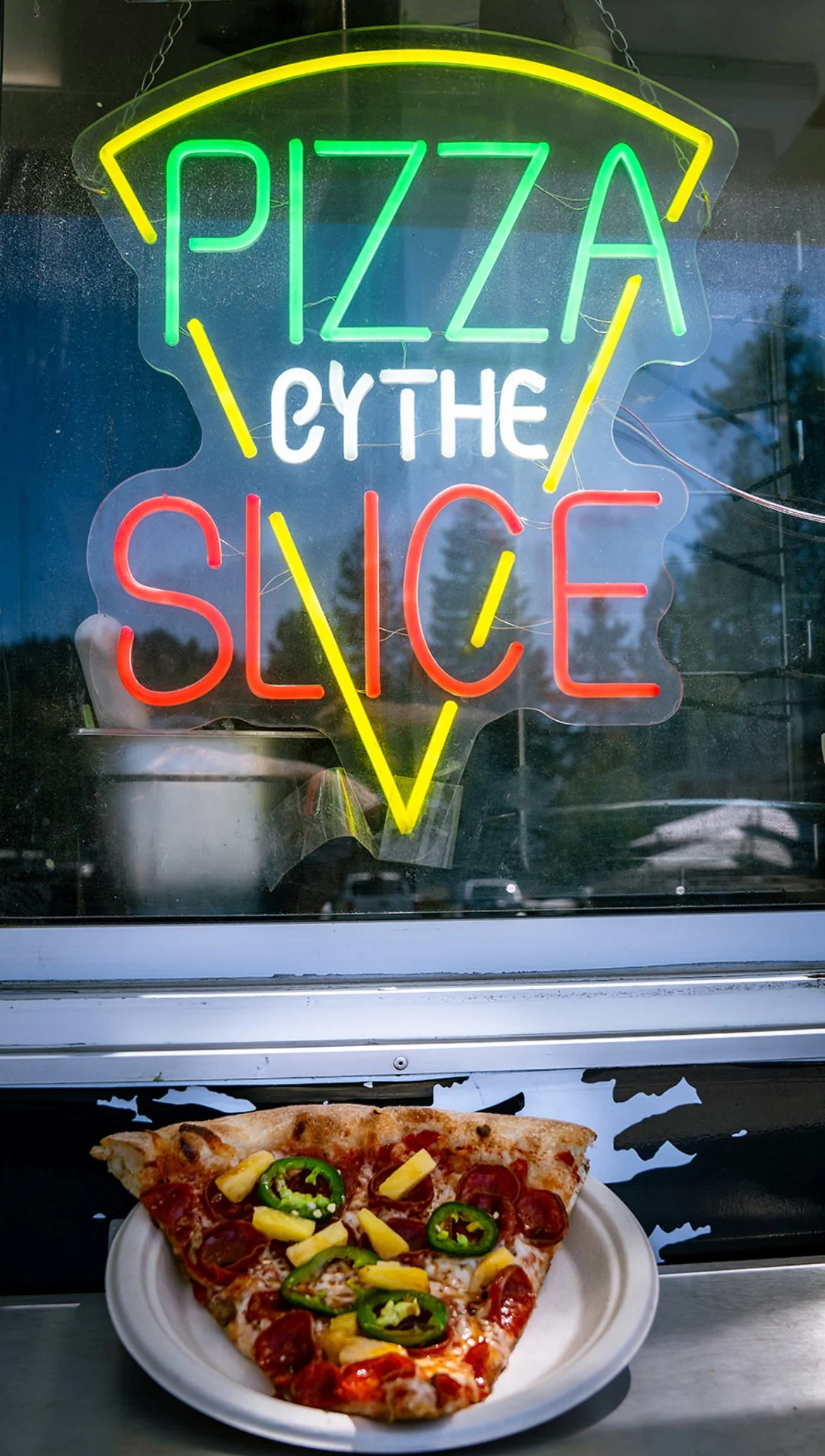 Neon sign reading 'Pizza Cythe the Slice' with a slice of pepperoni pizza topped with jalapeños and pineapple on a white plate in front of the sign.