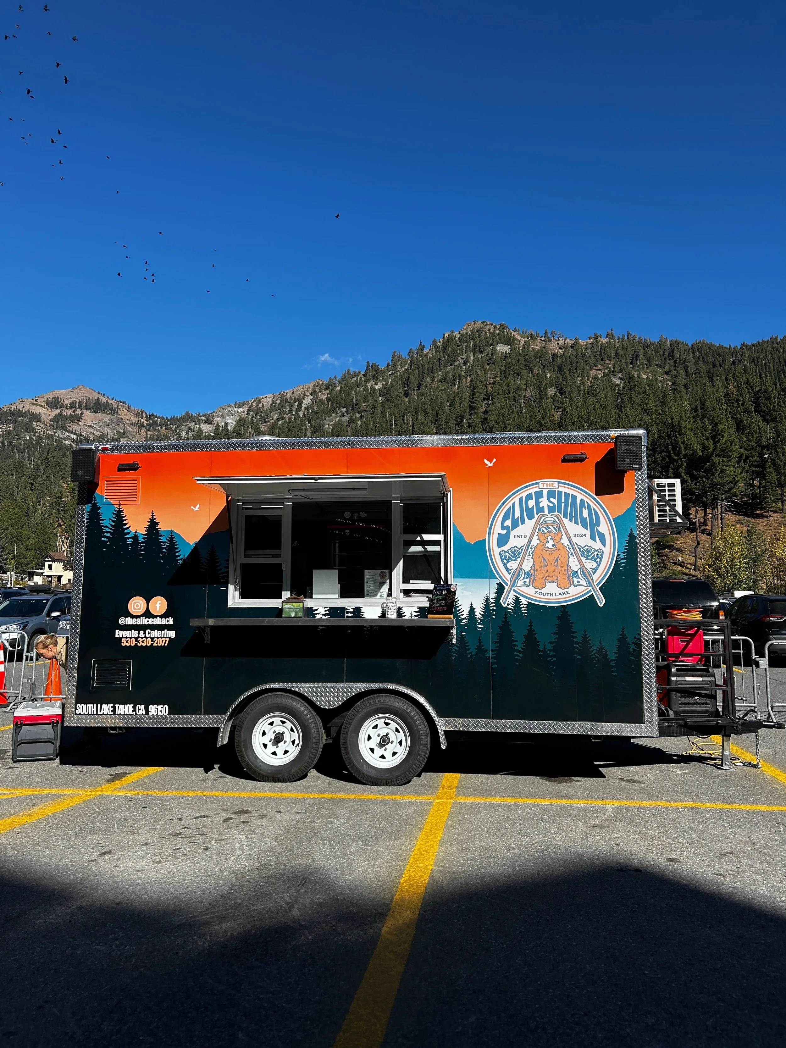 A food truck with mountain and forest graphics, parked in a lot with mountains and clear blue sky in the background.