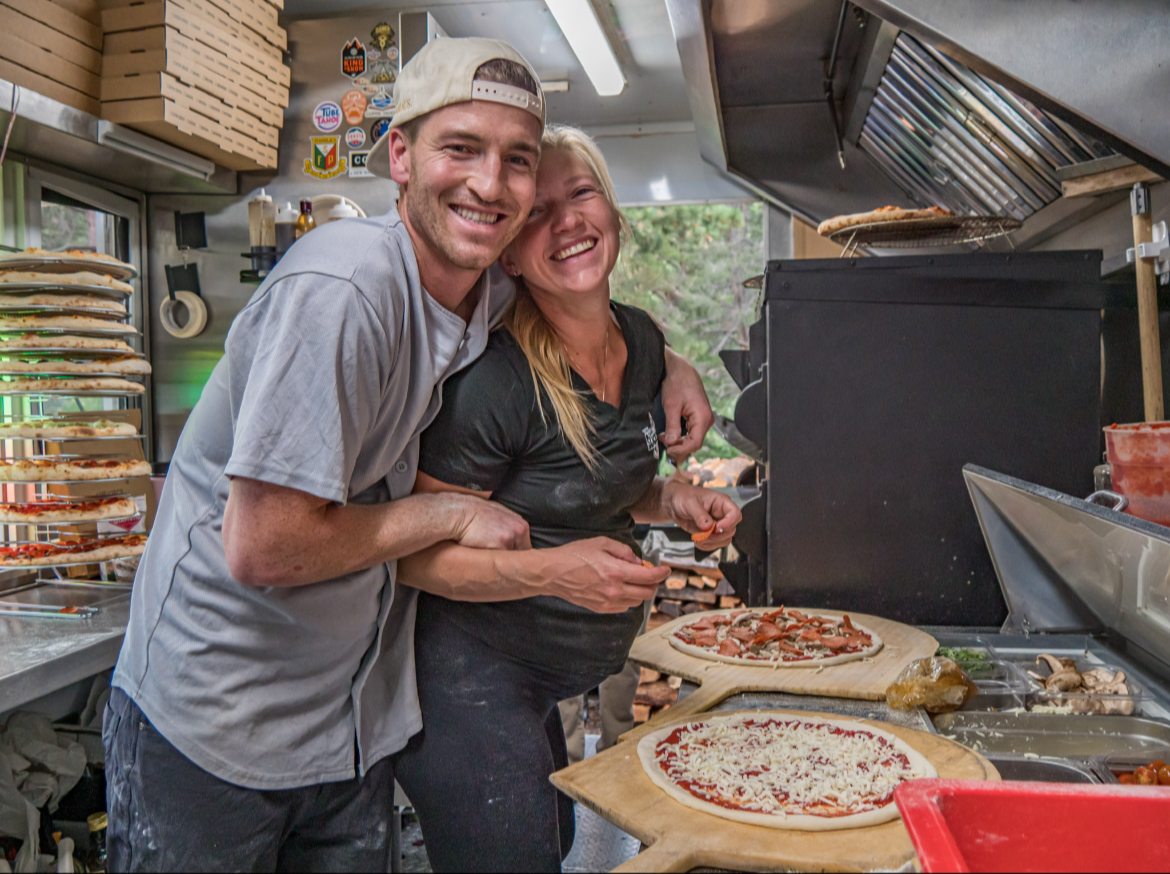 A man and a woman smiling inside a pizza food truck, preparing pizzas with toppings, surrounded by pizza supplies and kitchen equipment.