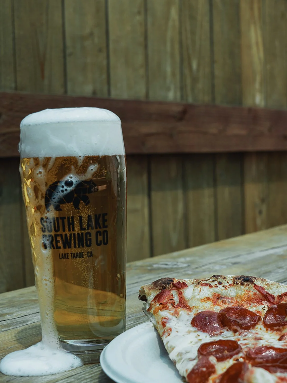 A glass of beer with foam on top and a slice of pepperoni pizza on a white plate on a wooden table, with a wooden fence in the background