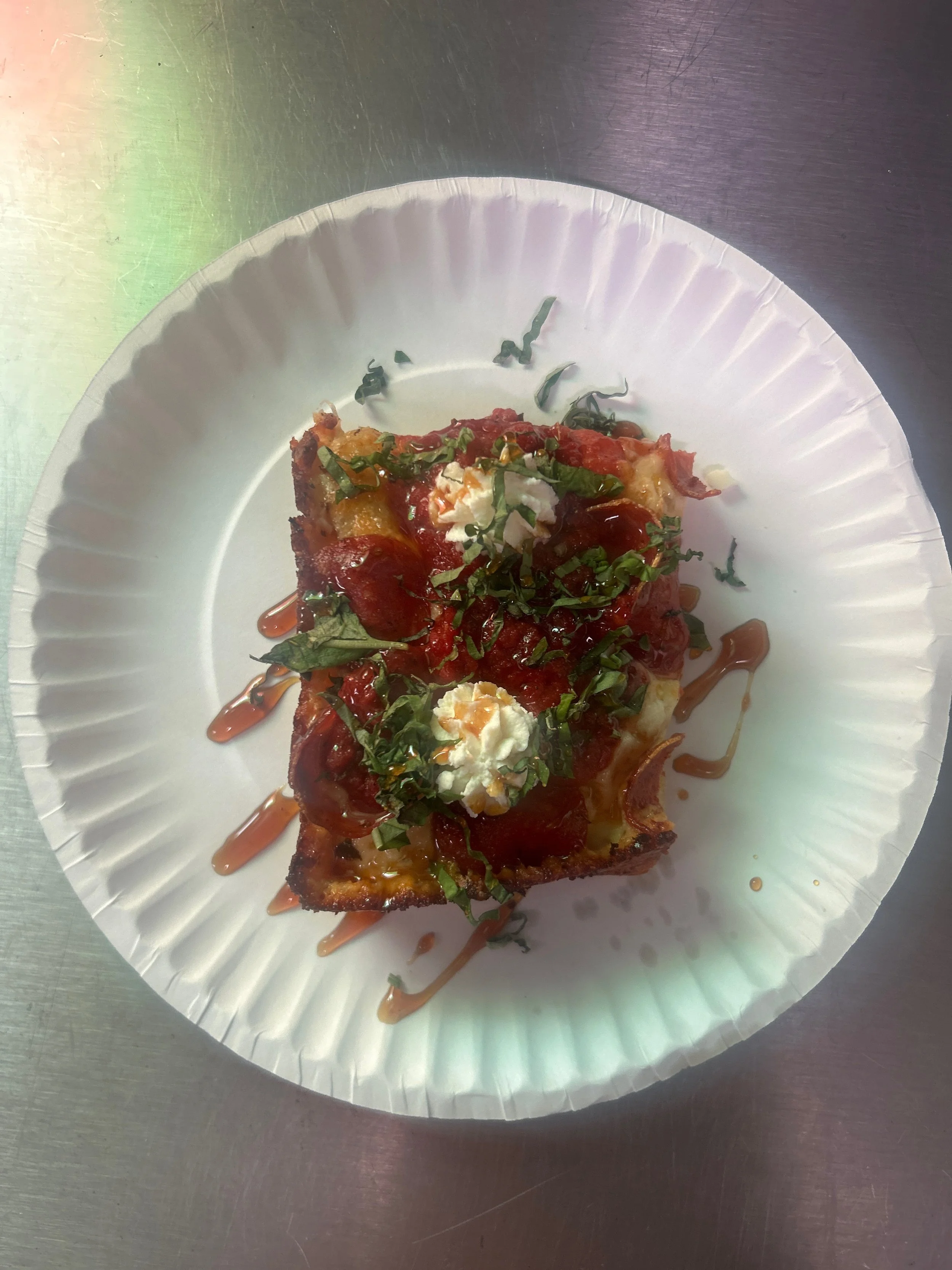 A slice of pizza topped with cheese, tomato sauce, chopped herbs, shredded cheese, and drizzled with a sauce, served on a white paper plate on a metal surface.