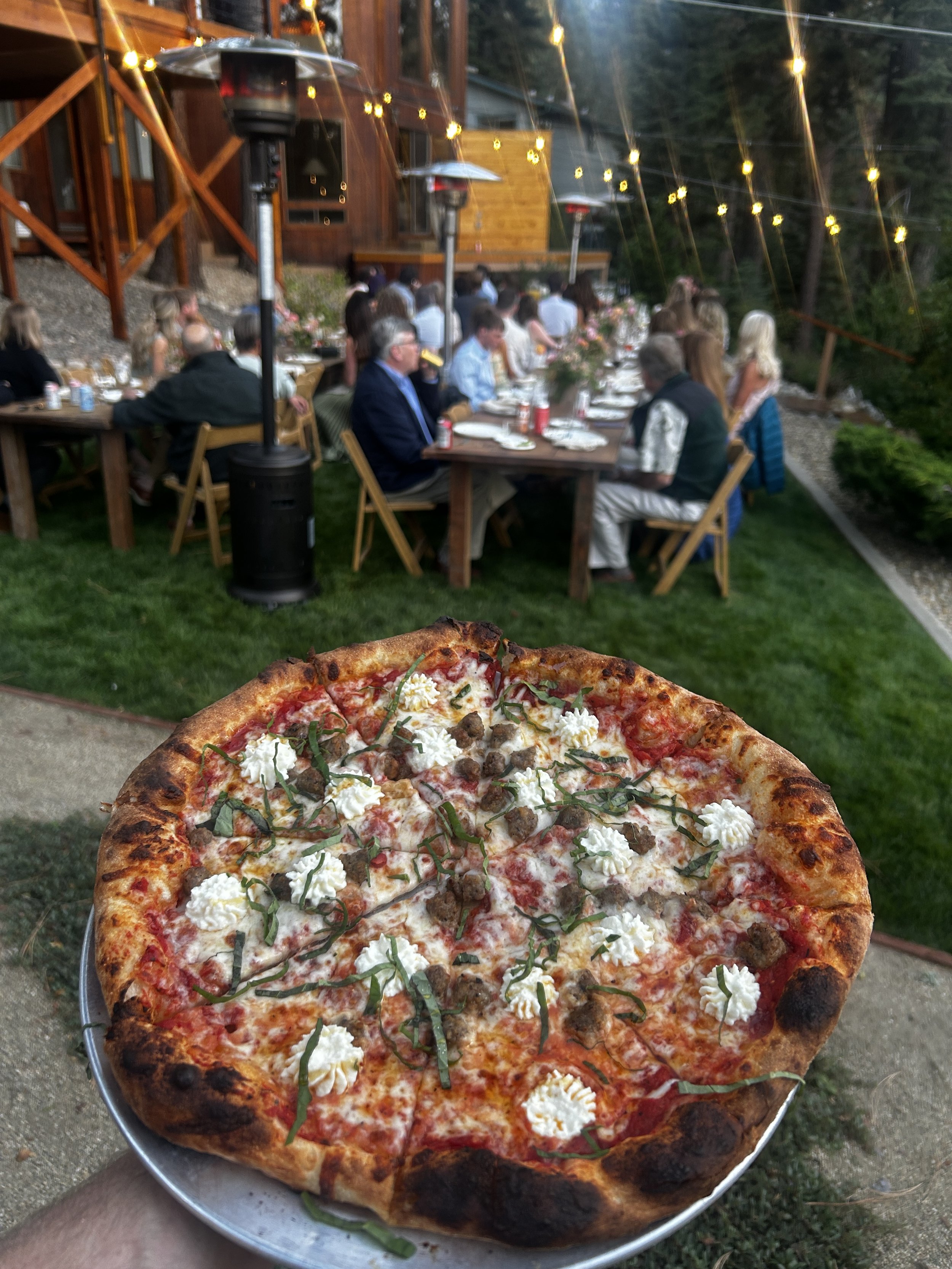 Close-up of a pizza with cheese, meatballs, and green peppers held outdoors, with a blurred outdoor dining area in the background.