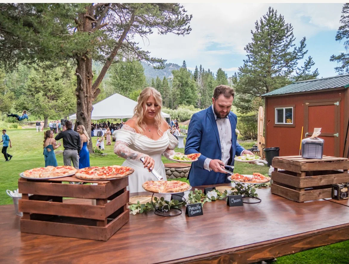 Bride and groom serving themselves pizza at an outdoor wedding reception in a grassy, wooded area with guests in the background.
