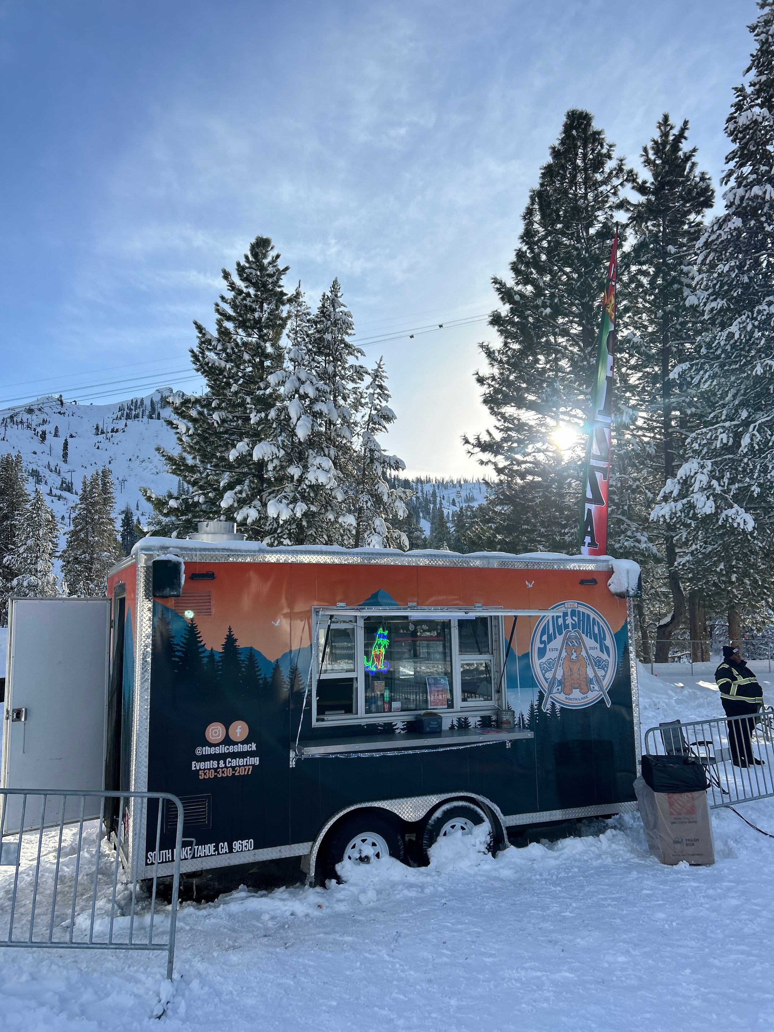 A ski shack food truck with a mountain and snow-covered trees in the background, set in a snowy landscape with a blue sky and sunlight peeking through the trees.