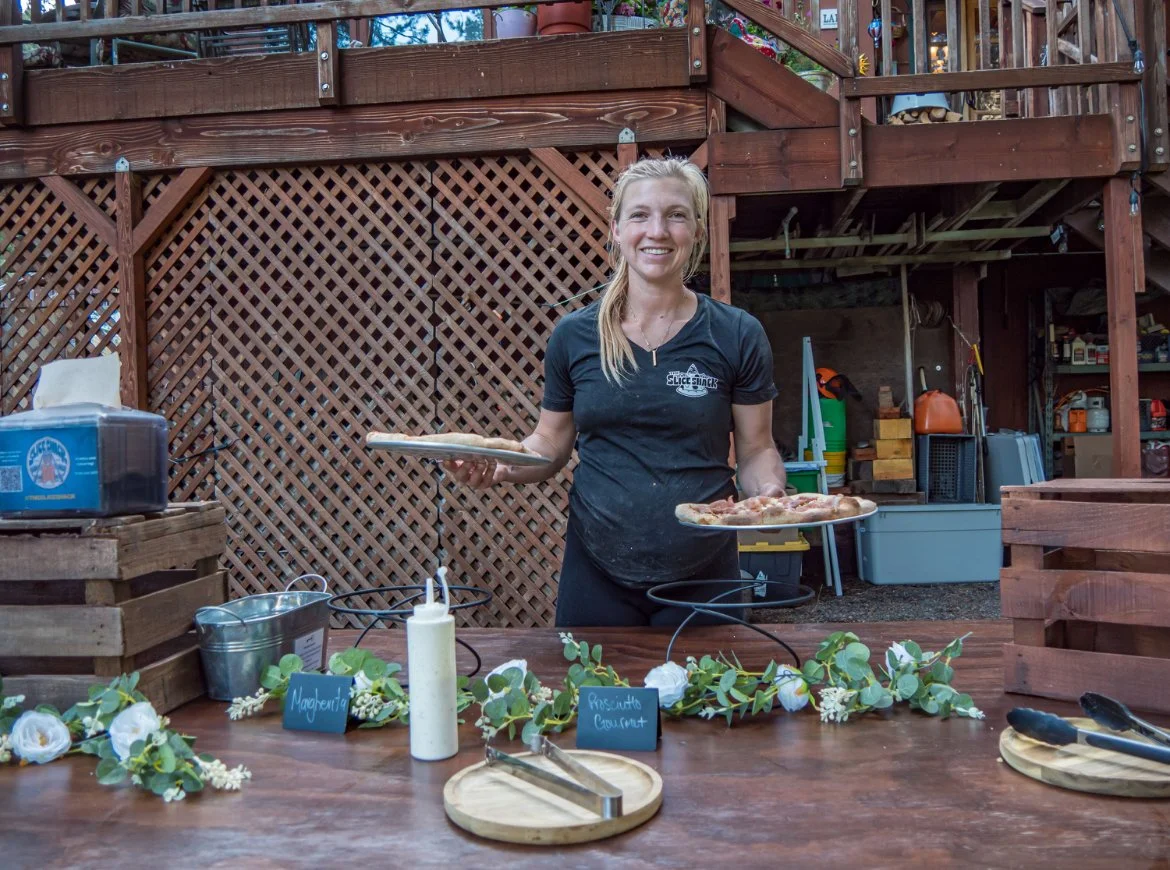A woman standing behind a table in a backyard, holding pizza slices on plates. The table is decorated with green foliage and white flowers, with containers, tongs, and a bottle on it.