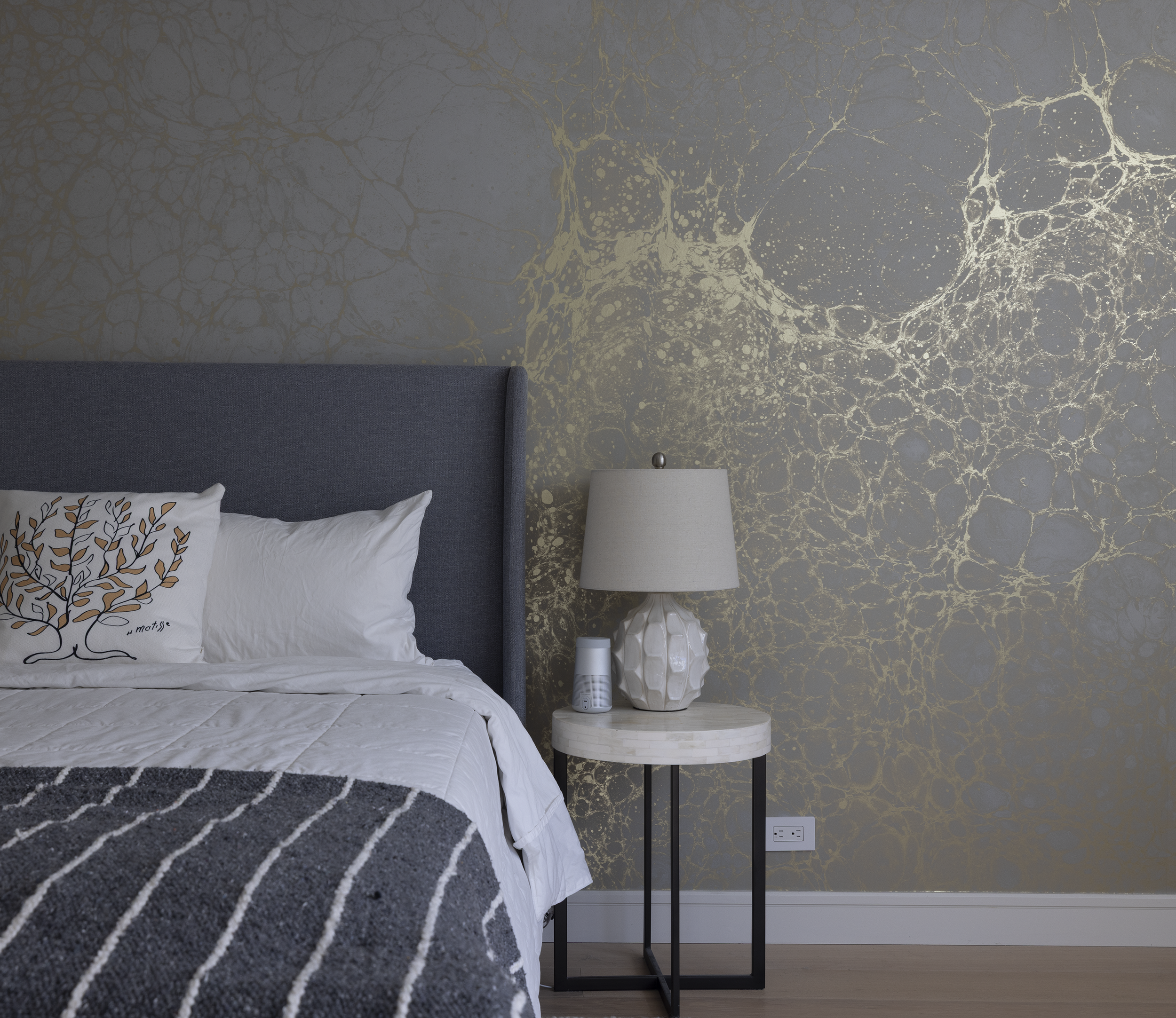 A bedroom with a dark gray upholstered headboard, a bed with white and gray bedding, a decorative pillow with a tree, a white textured table lamp on a small round side table, and patterned wallpaper with gold accents behind the lamp.