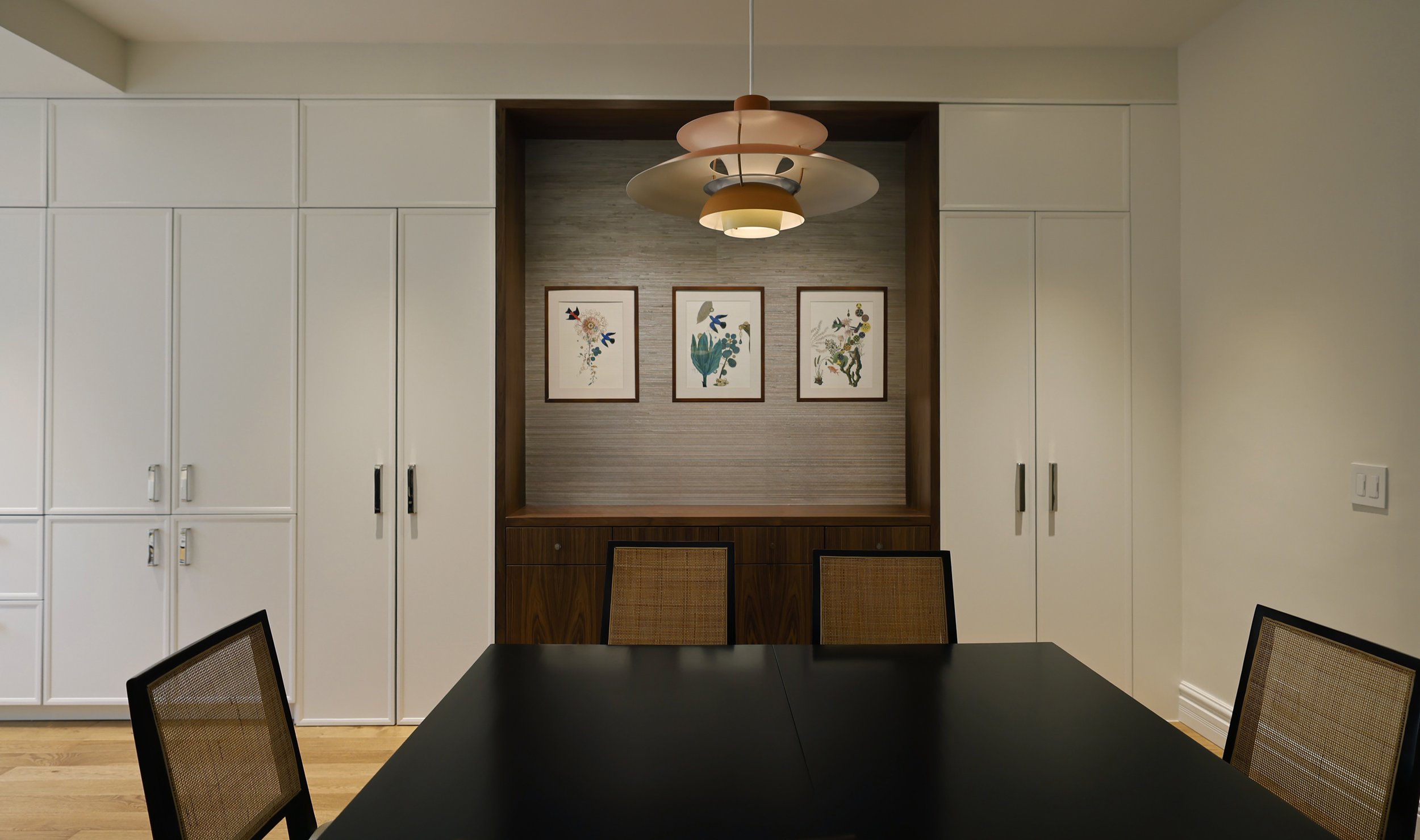 Dining room with black table, woven chairs, built-in white cabinets, and a sideboard with decorative niche covered in grasscloth with botanical art prints and a layered pendant light.