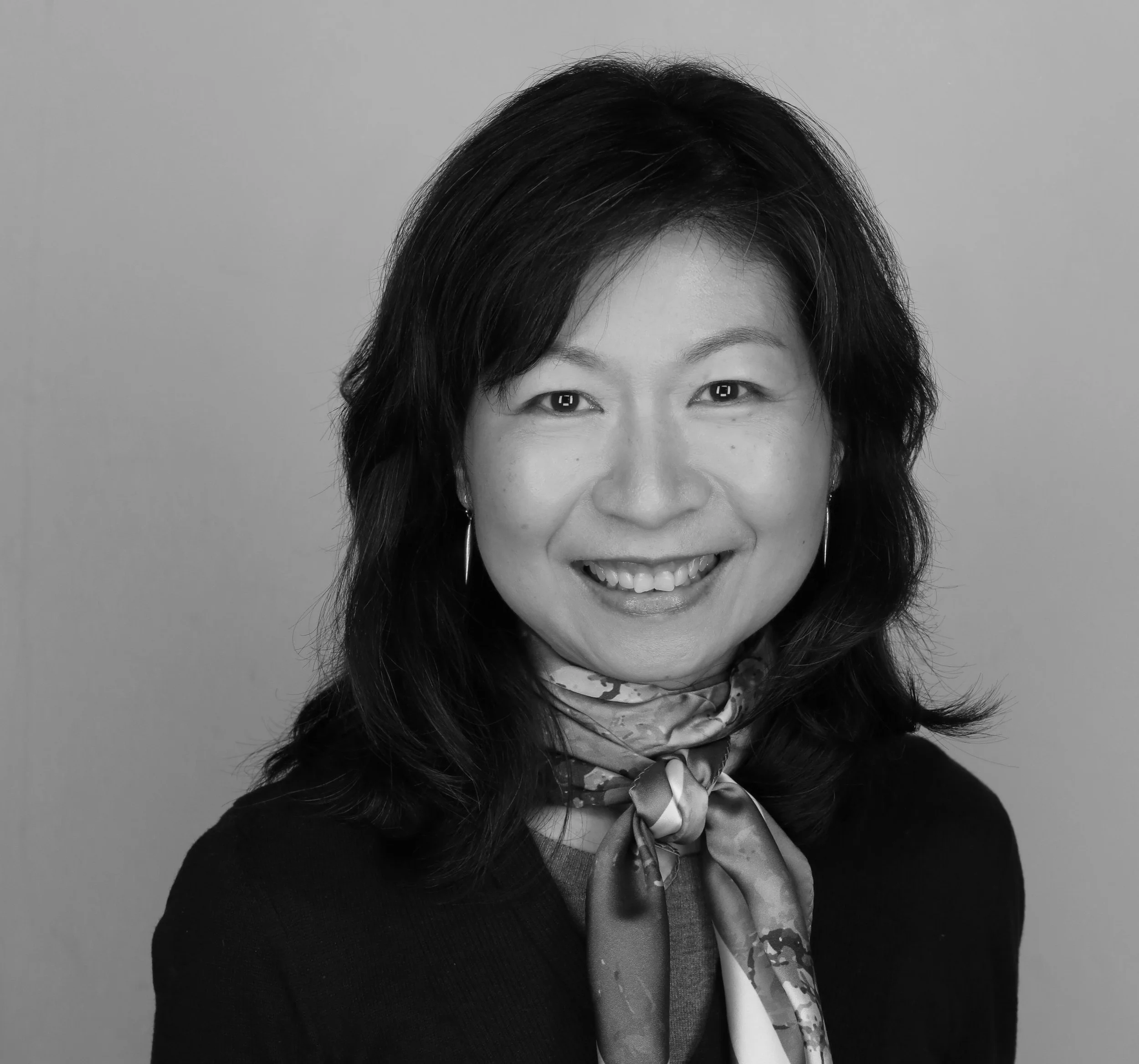 Partial black and white portrait of a woman with dark hair, smiling, wearing earrings, a patterned scarf, and a dark top.