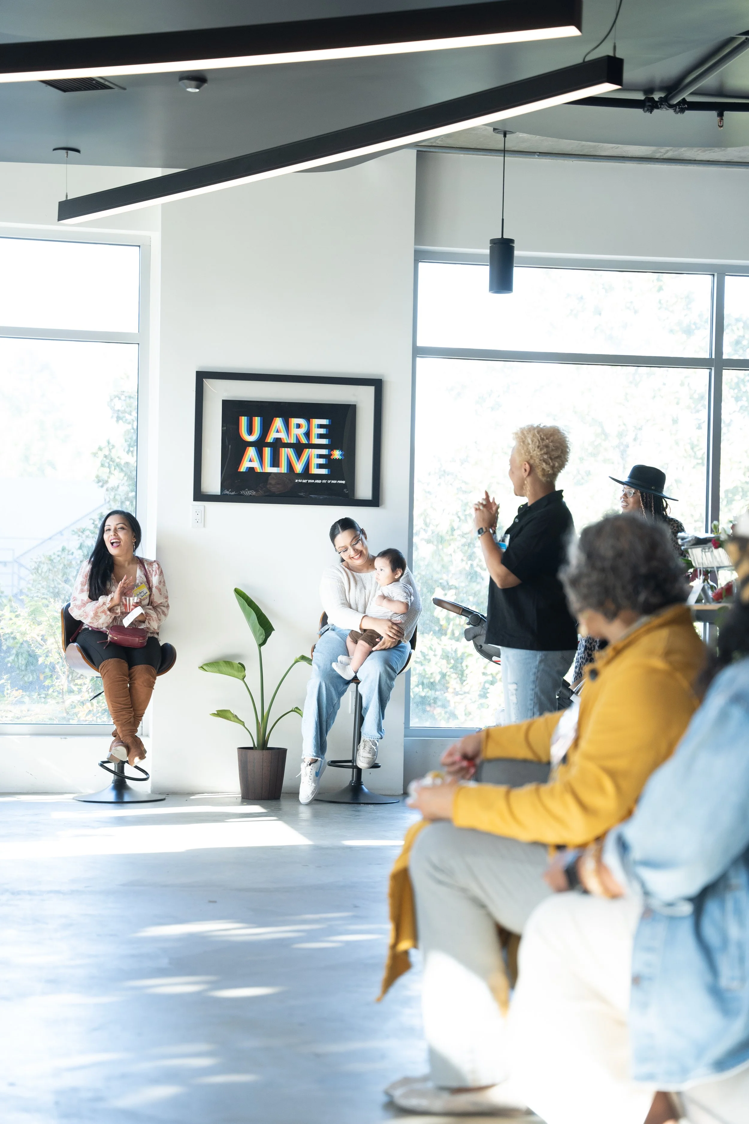 Group of diverse women and a child in a modern, brightly lit room with large windows, some sitting on chairs and one standing, engaging in a conversation or presentation; a framed sign on the wall reads 'U ARE ALIVE' in colorful text.